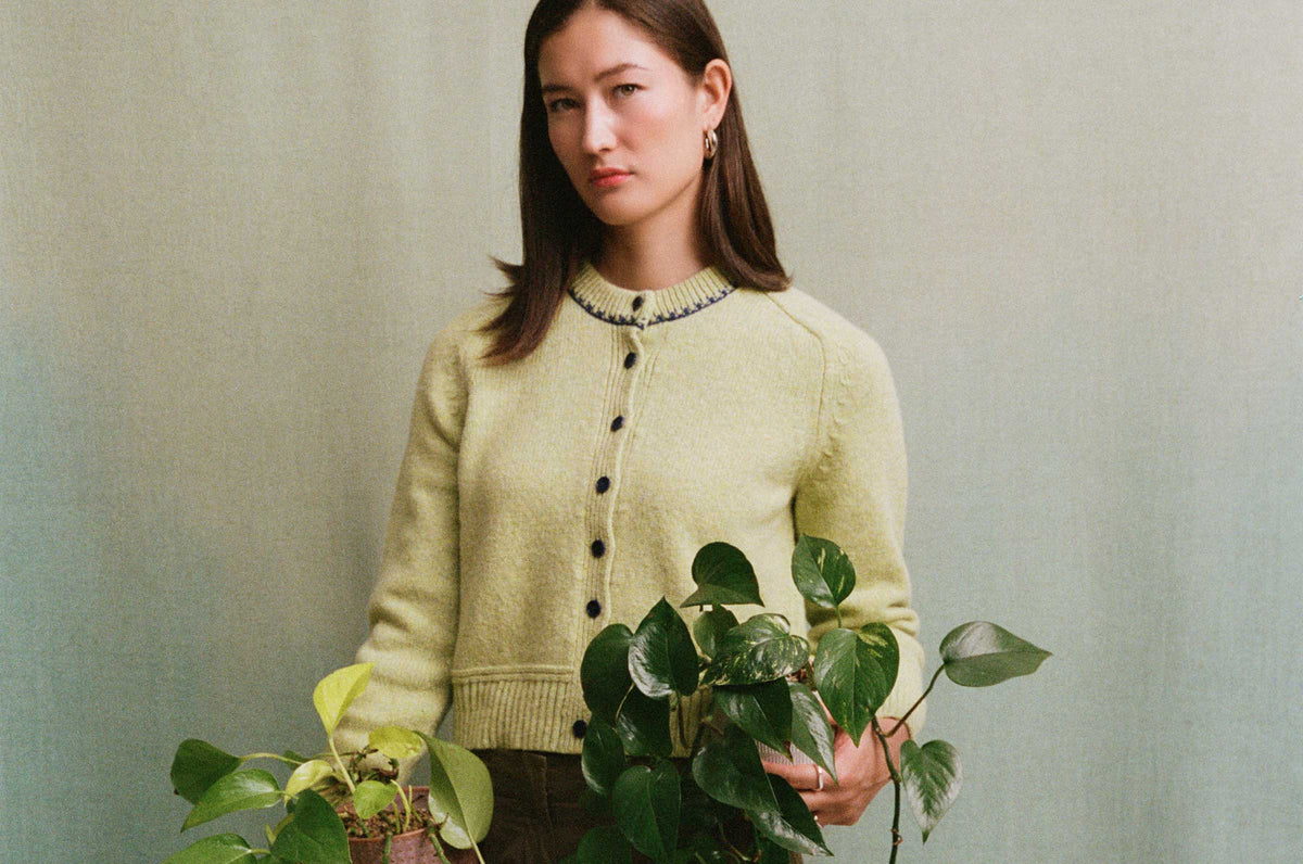 Lime Cecilia cardigan with navy buttons and contrast neckline stitching, worn by a model holding leafy plants against a soft green background.