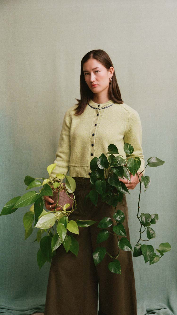 Lime Cecilia cardigan with navy buttons and contrast neckline stitching, worn by a model holding leafy plants against a soft green background.
