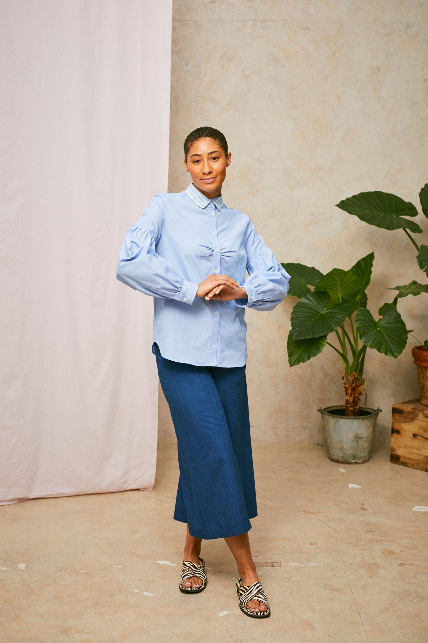 Model wears Saywood's pale blue shirt, the Edi Volume Sleeve Shirt with lace collar, with the natural indigo Japanese denim Amelia wide leg trousers and zebra print sandals. A plant and drop of pink fabric can be seen in the background. 