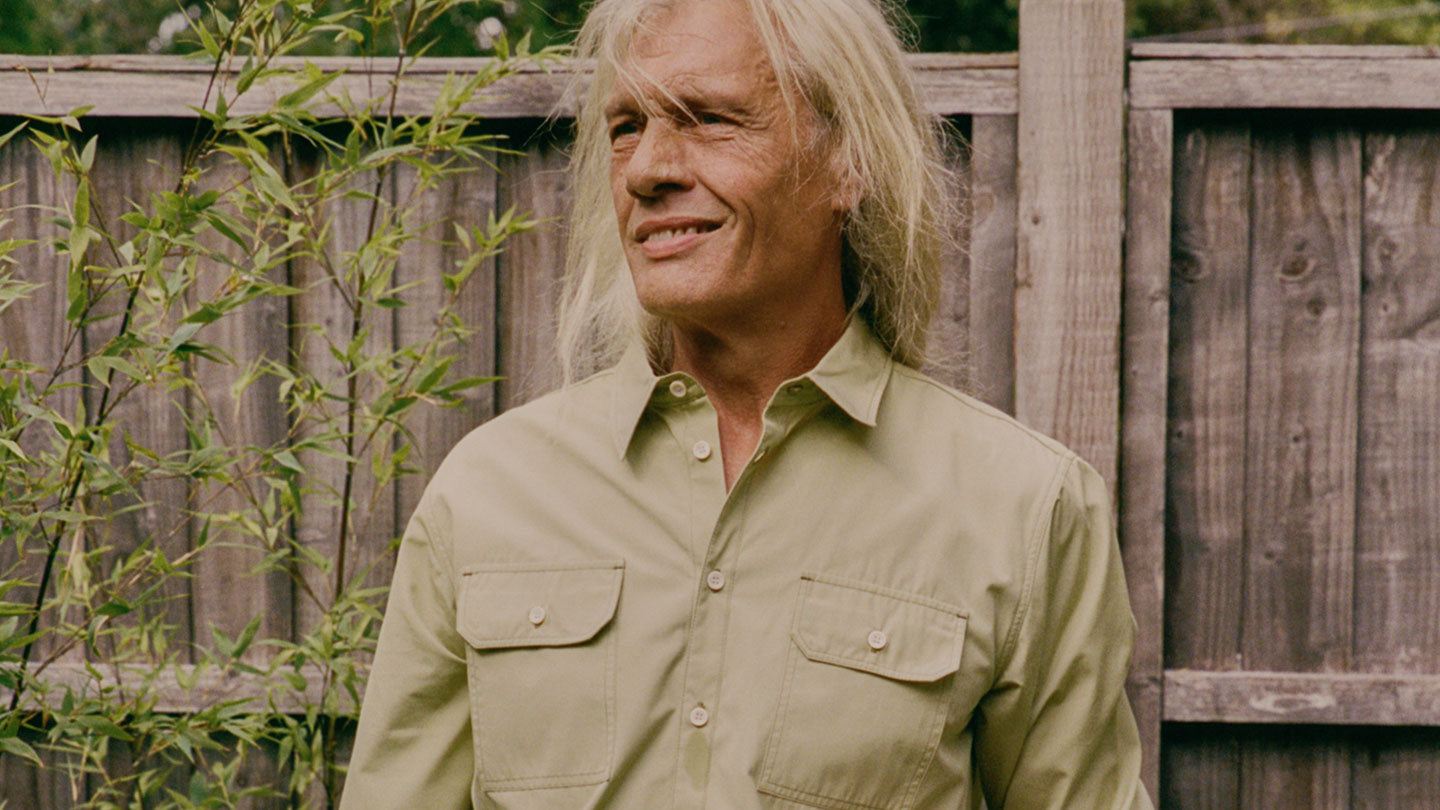 A man with long blonde grey hair stands in the garden in front of a plant bed and a wooden fence. He wears Saywood's Men's Eddy Utility Shirt with utility style pockets in olive.