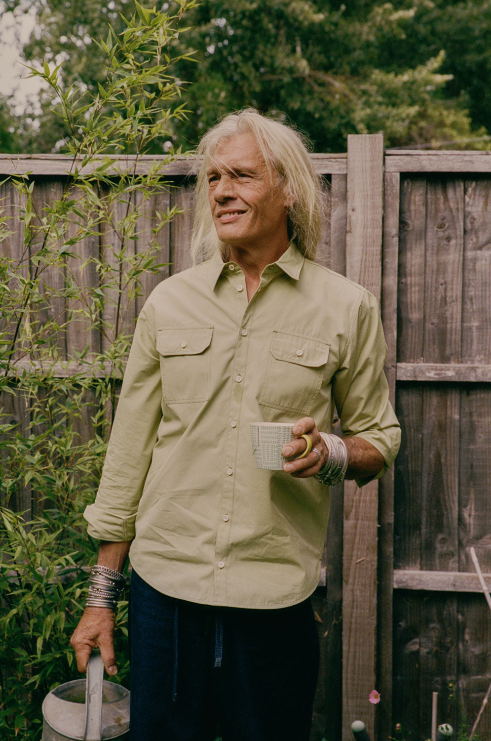 Man holding a mug outdoors, with a wooden fence and plants in the background. He is holding a watering can, and is wearing Saywood's men's Eddy Utility Shirt with utility style pockets in khaki olive.