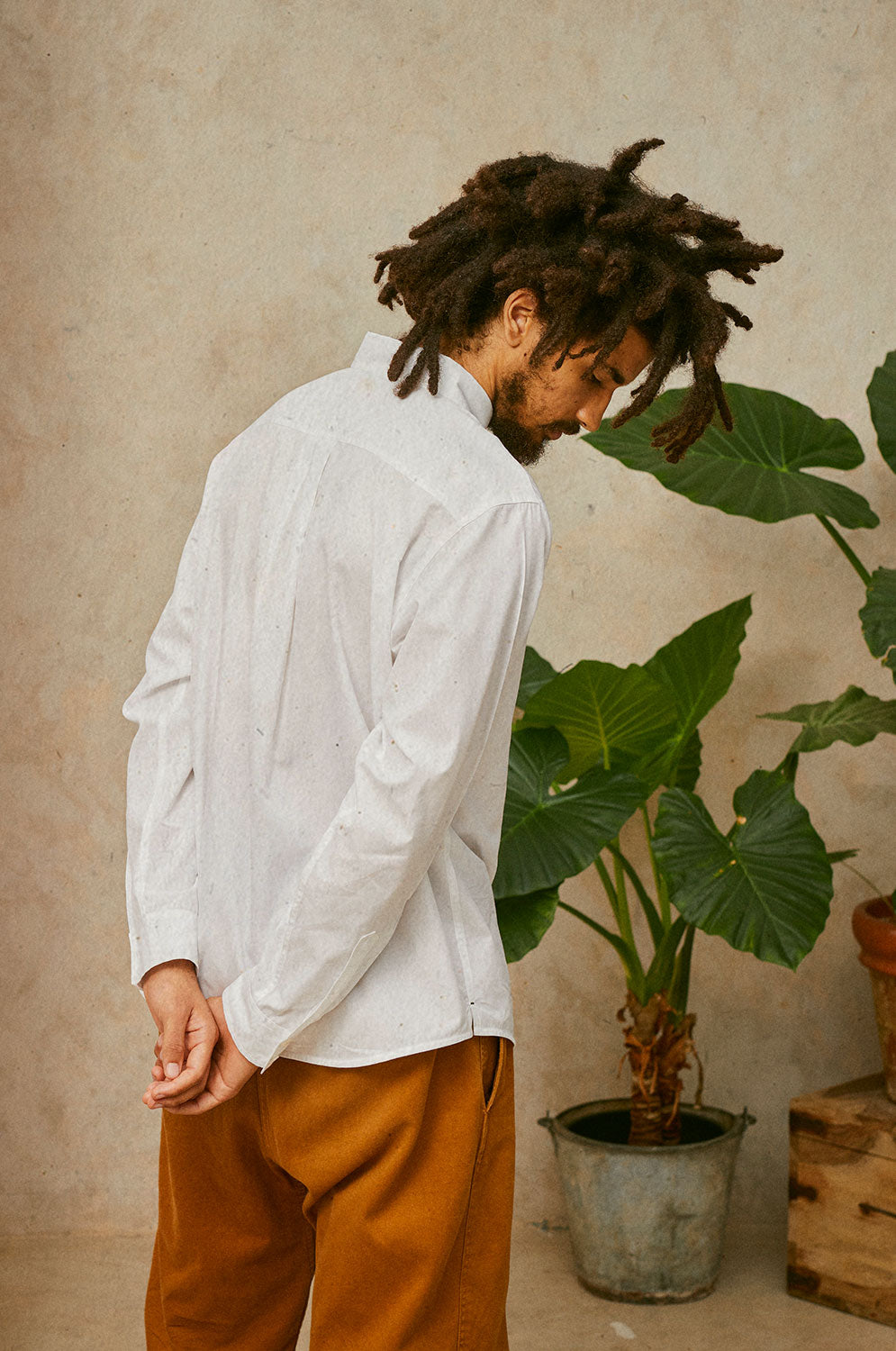 Back view of a man with locks wearing Saywood's Eddy Mens Classic White Shirt. Worn with tobacco trousers just visible. Model has both hands behind his back and looks towards the floor. A plant can be seen in the background.