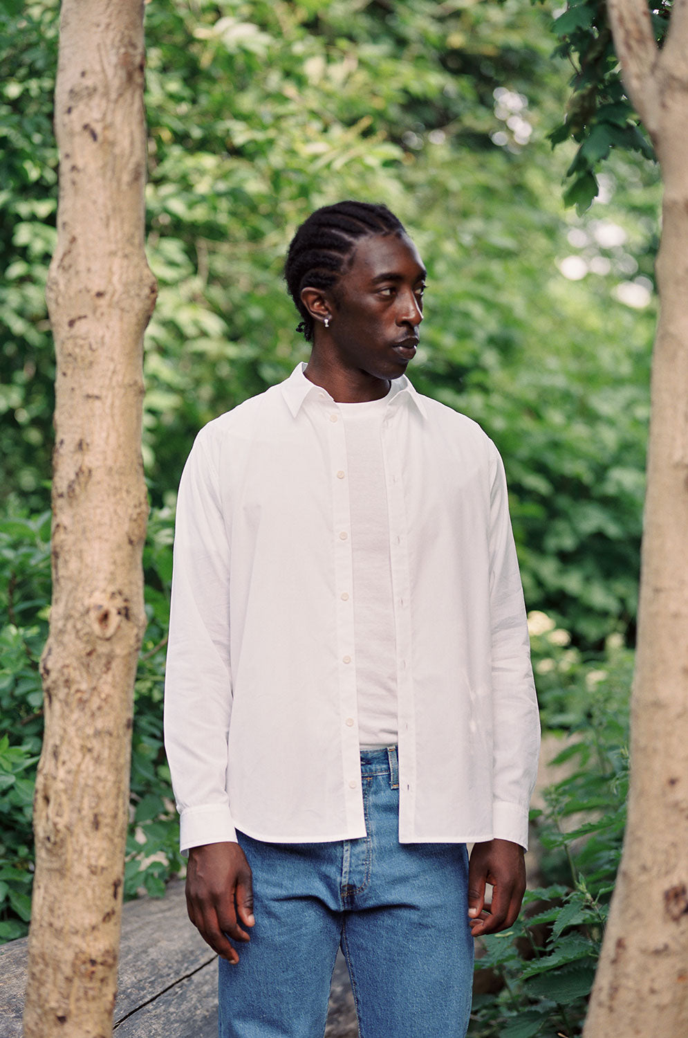 A black man is standing amongst the trees, looking to the side. He is wearing Saywood's Eddy Men's Classic White Shirt with long sleeves, worn open over a white t-shirt and jeans.