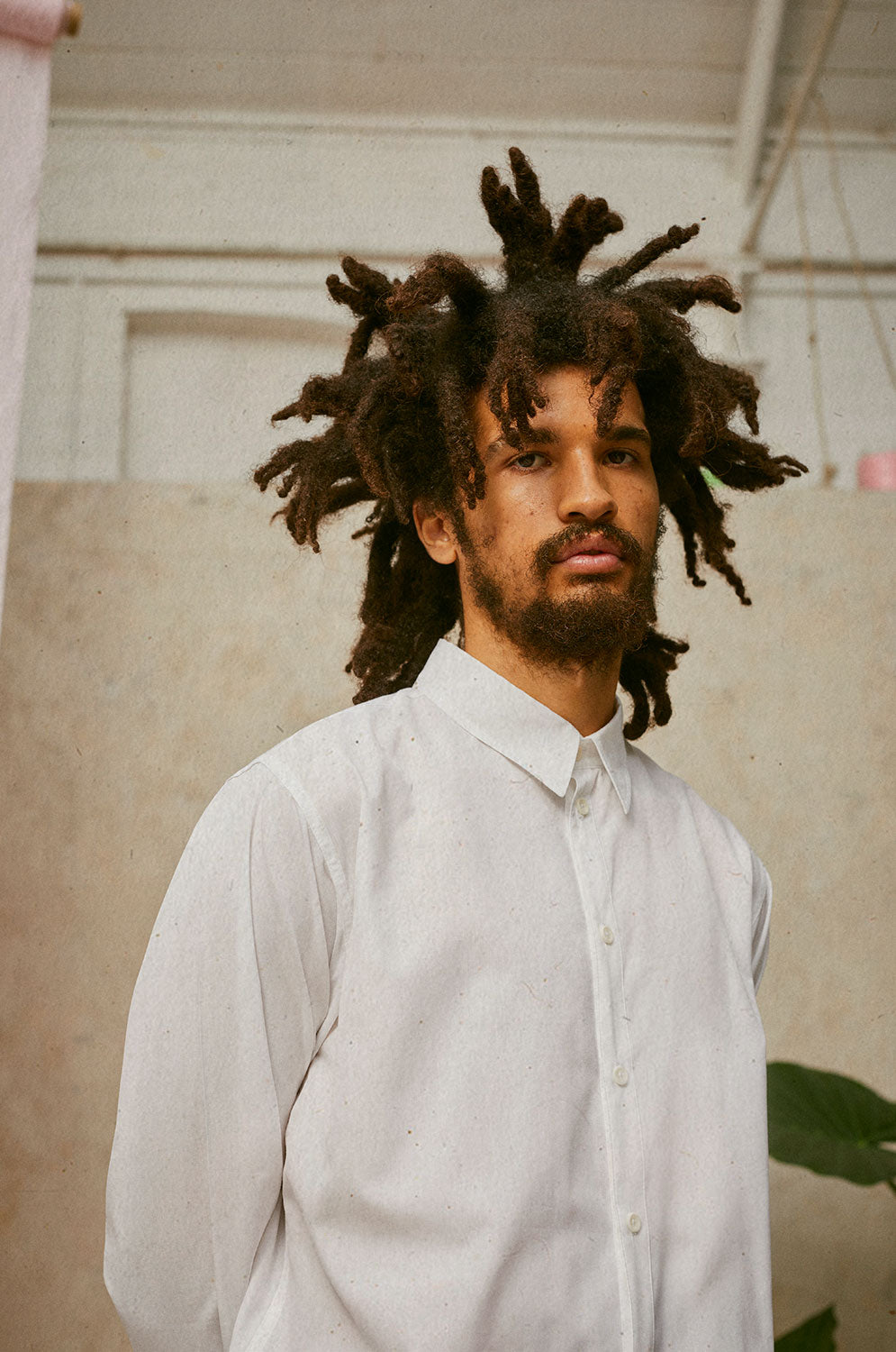 A man with locks is wearing Saywood's Eddy Mens Classic White Shirt. he has both hands behind his back. The collar and corozo button placket can be seen.