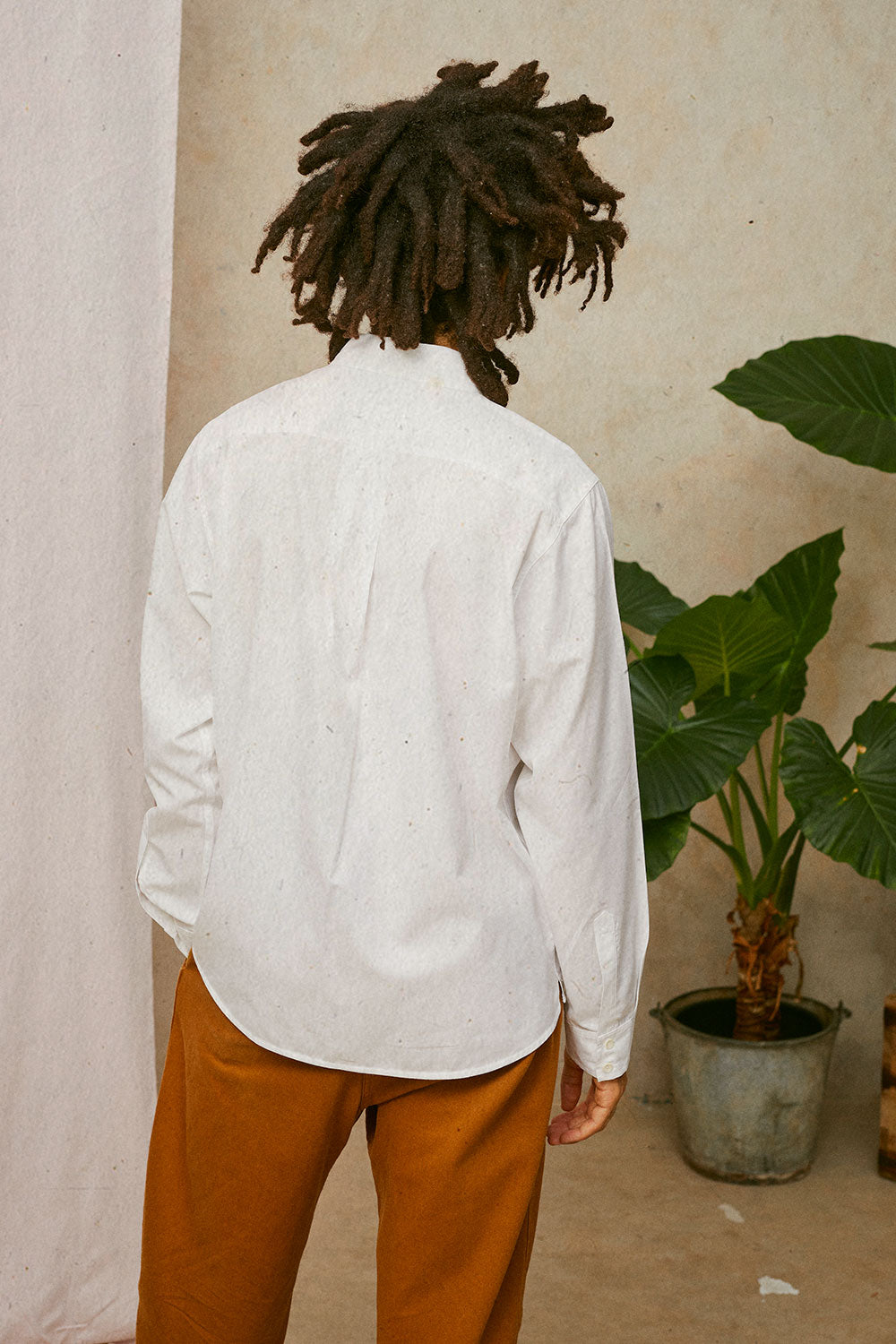 Back view of a man with locks wearing Saywood's Eddy Mens white Shirt with patch pockets; showing the back yoke and box pleat in the back. Worn with tobacco trousers. Model has one hand in his trouser pocket. A plant and drop of pink fabric can be seen in the background.
