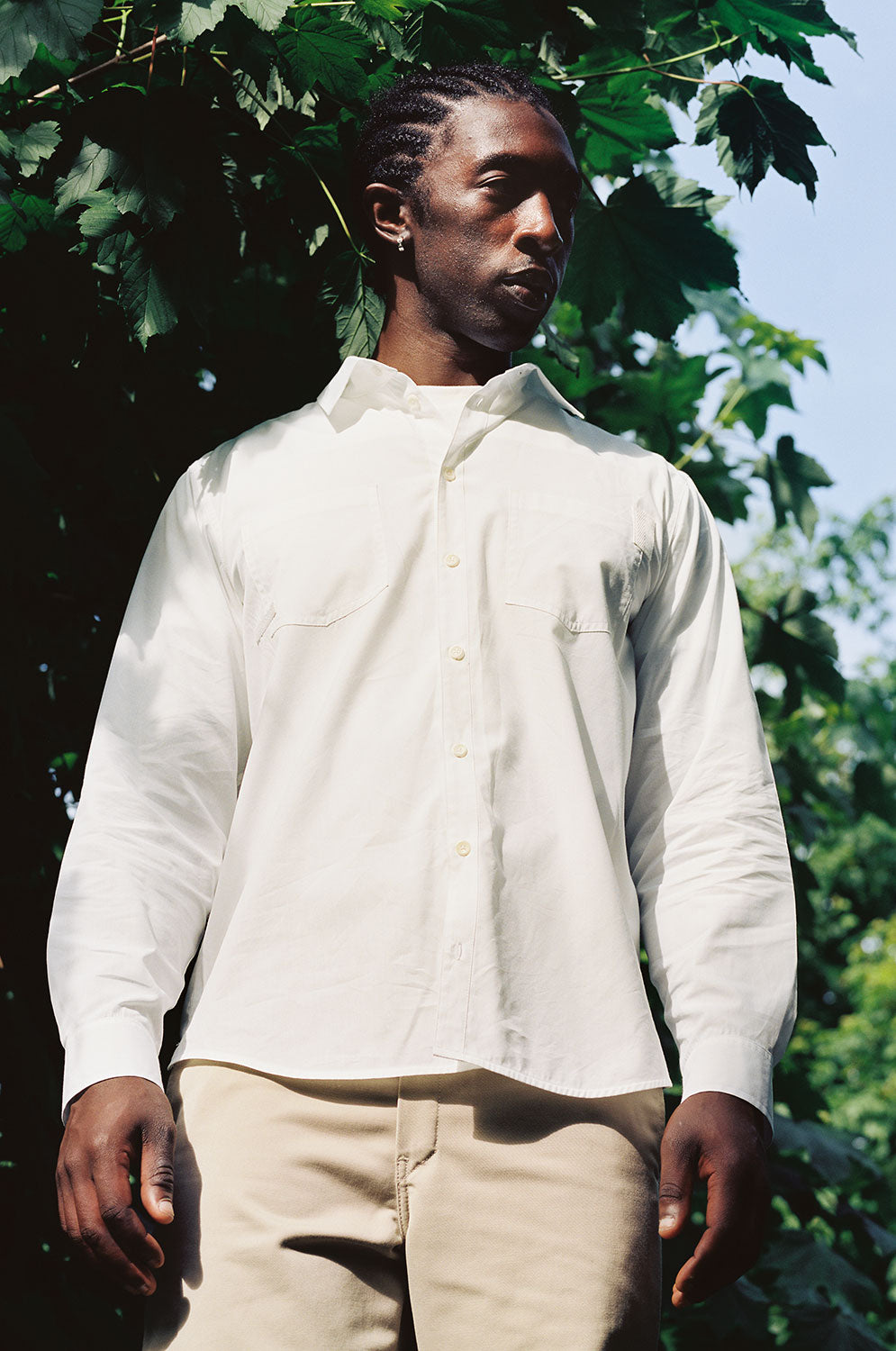 Close up of a black man wearing Saywood's Eddy Mens white Shirt with patch pockets, worn with the top buttons open and a white t-shirt underneath. The model is surrounded by trees and greenery.