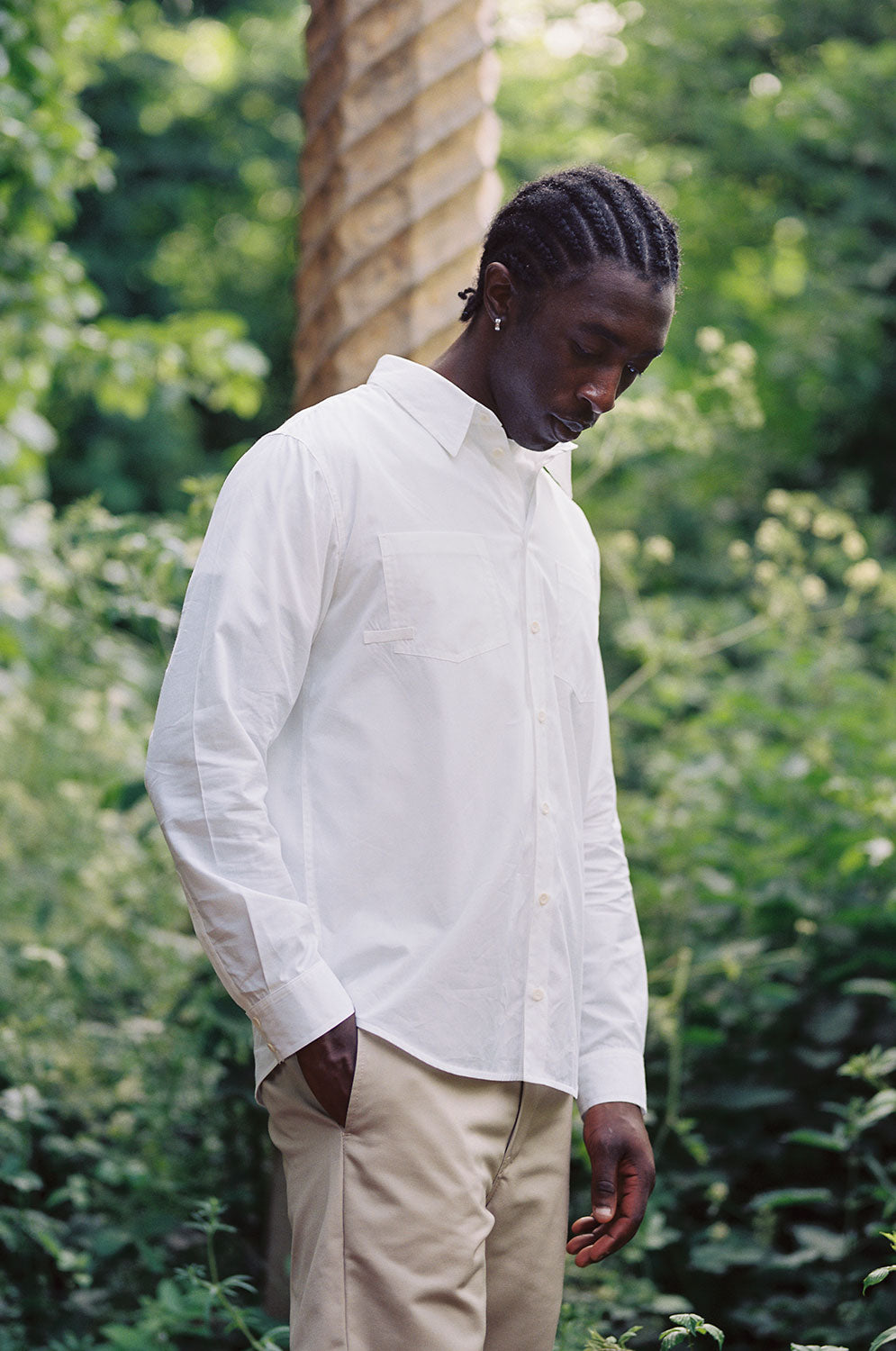 A black man wearing Saywood's Eddy Mens white Shirt with patch pockets, worn with the top buttons open and a white t-shirt underneath. He is surrounded by trees and greenery, with one hand in his trouser pocket and looking down.