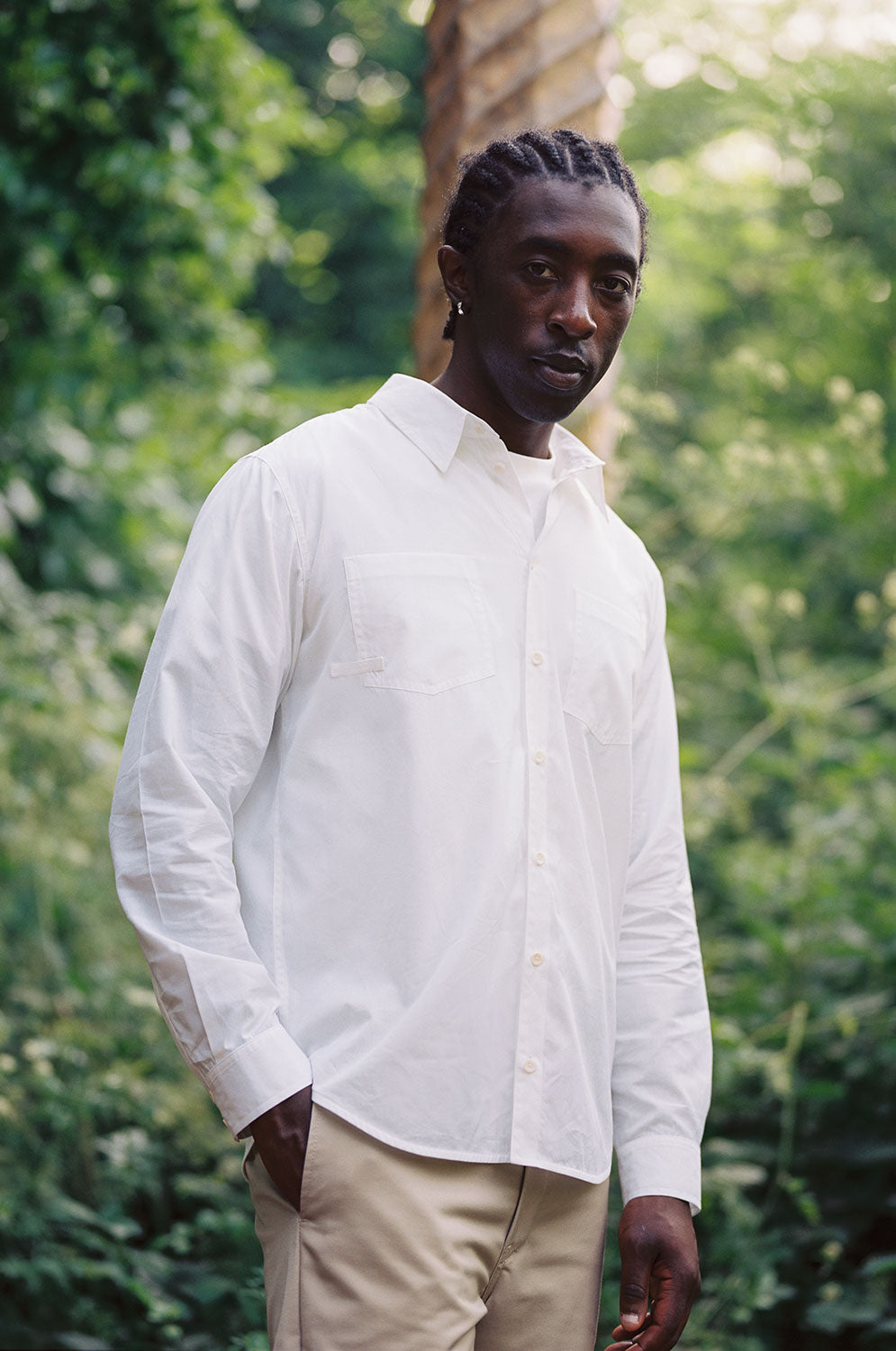 Close up of a black man wearing Saywood's Eddy Mens white Shirt with patch pockets, worn with the top buttons open and a white t-shirt underneath. He is surrounded by trees and greenery, with one hand in his trouser pocket and looking at the camera.