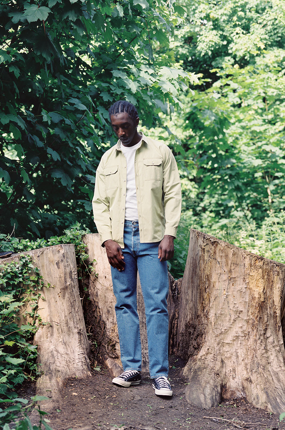 A man stands in front of several large tree stumps, surrounded by greenery. He wears Saywood's Eddy Mens Khaki Shirt with utility pockets, worn open with a white t-shirt underneath. Worn with jeans.