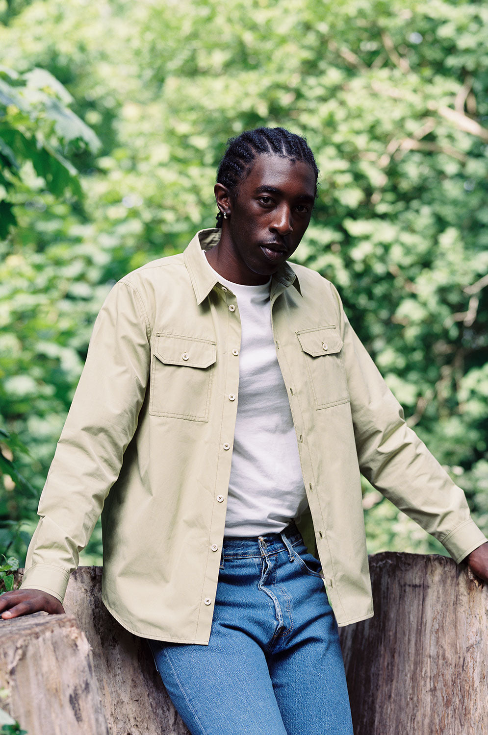 A man sits leaning on a large tree stump surrounded by greenery. He wears Saywood's Eddy Mens Khaki Shirt with utility pockets, worn open with a white t-shirt underneath. Worn with jeans.