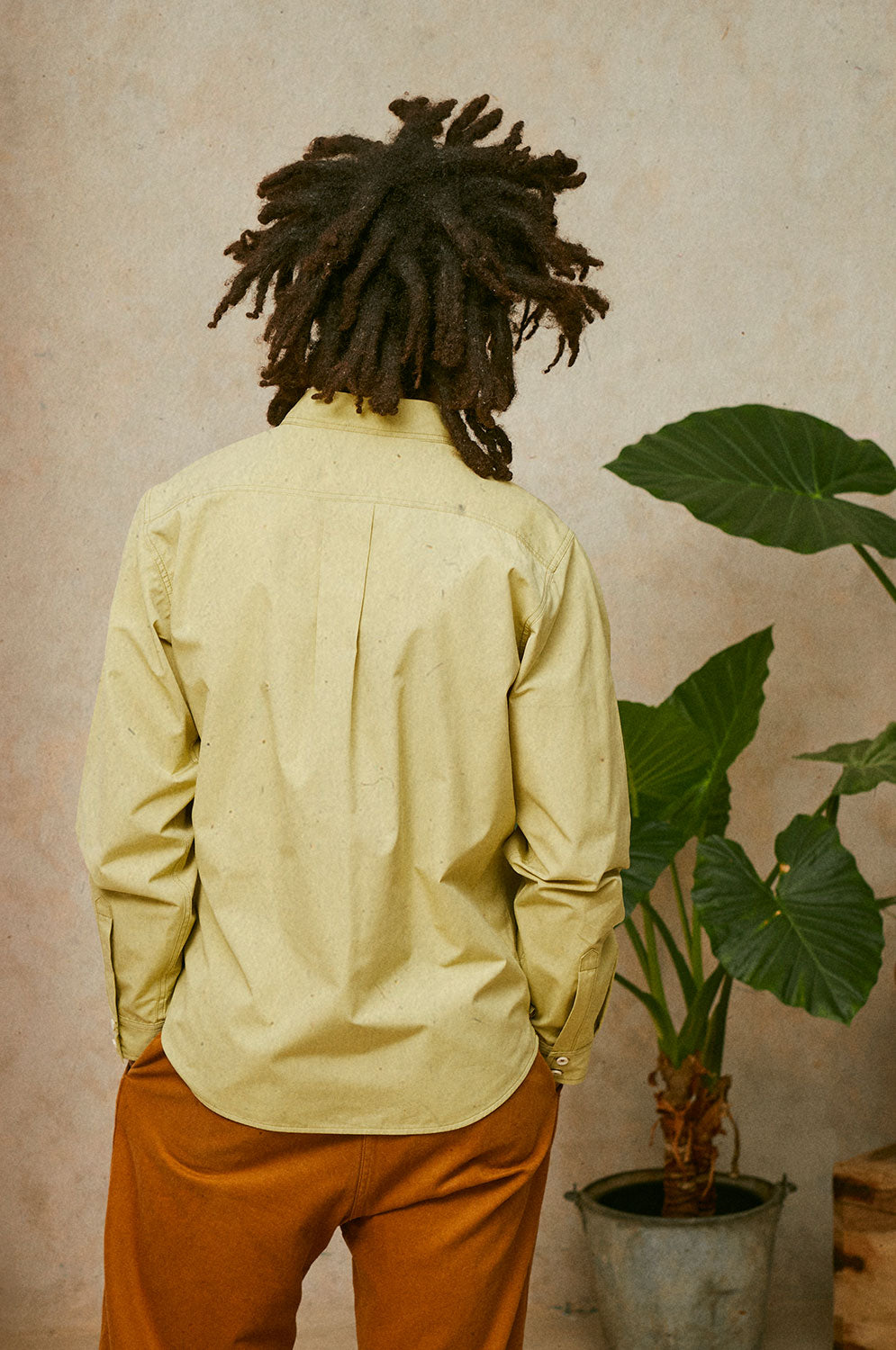 Close up back view of a man wearing Saywood's Eddy Mens Khaki Utility Shirt with long sleeves. Worn with tobacco trousers just visible. He has both hands in his trouser pockets and a plant can be seen in the background.