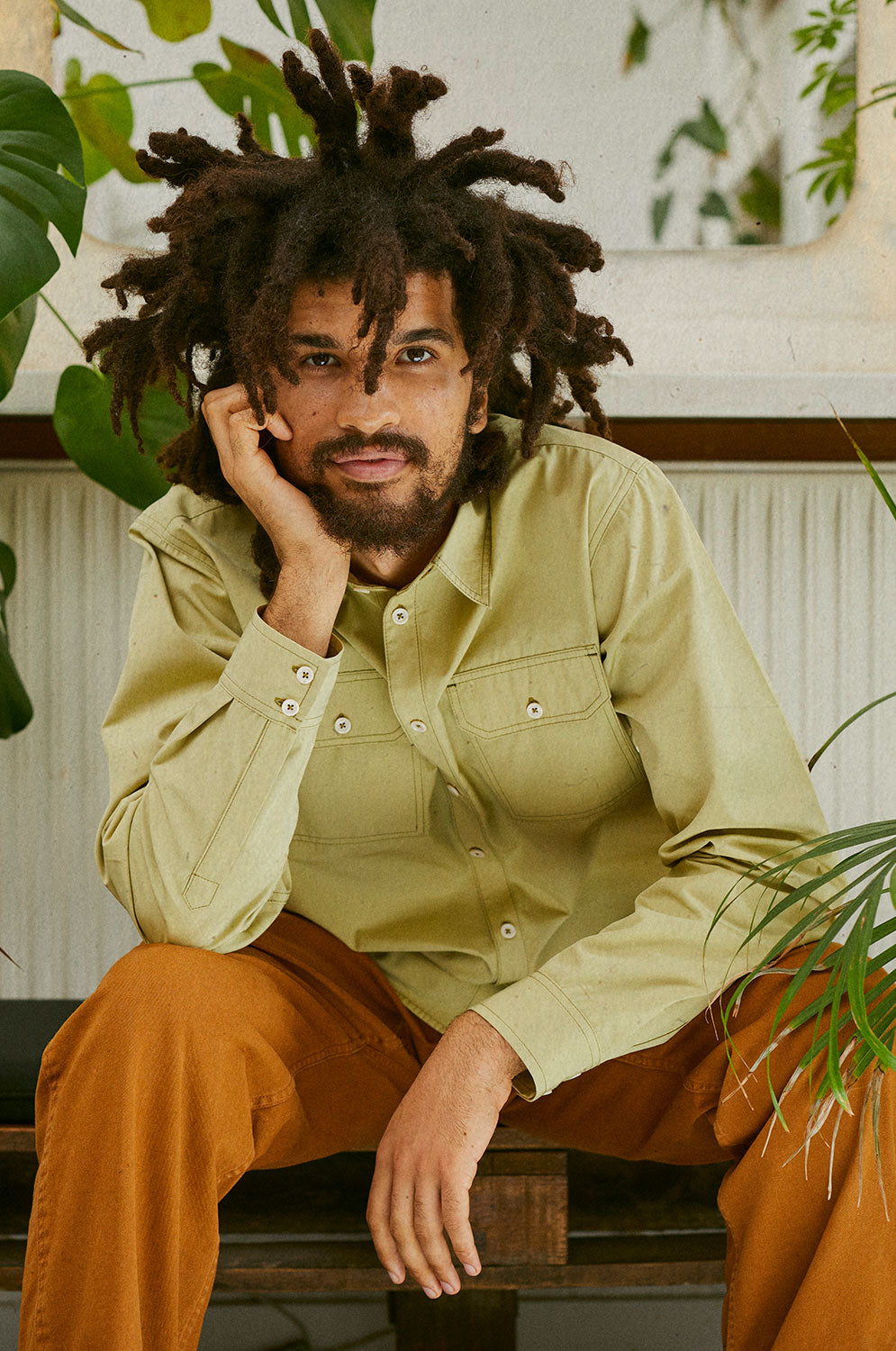 A man is sitting on a bench surrounded by house plants. One arm leans on his leg and his head leans on the other hand. He's wearing Saywood's Eddy Mens Khaki Shirt with utility pockets. Worn with tobacco trousers.
