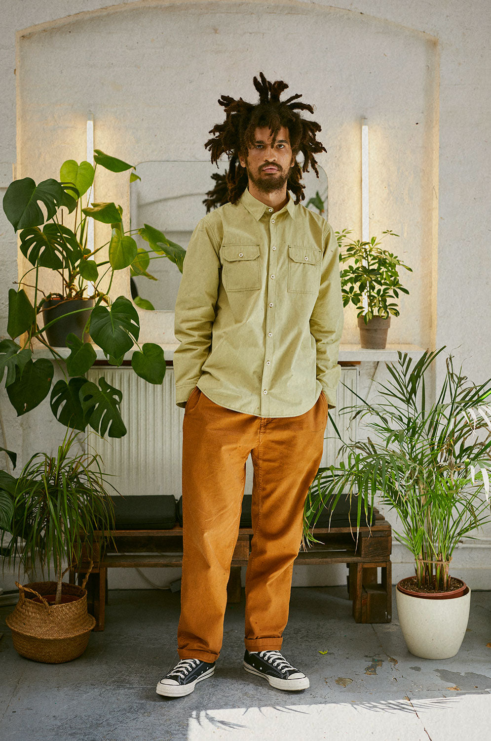 A man in front of a mirror and bench made of crates, surrounded by house plants. He has both hands in his pockets and is looking at the camera. He's wearing Saywood's Eddy Mens Khaki Utility Shirt with utility pockets and long sleeves. Worn with tobacco trousers.
