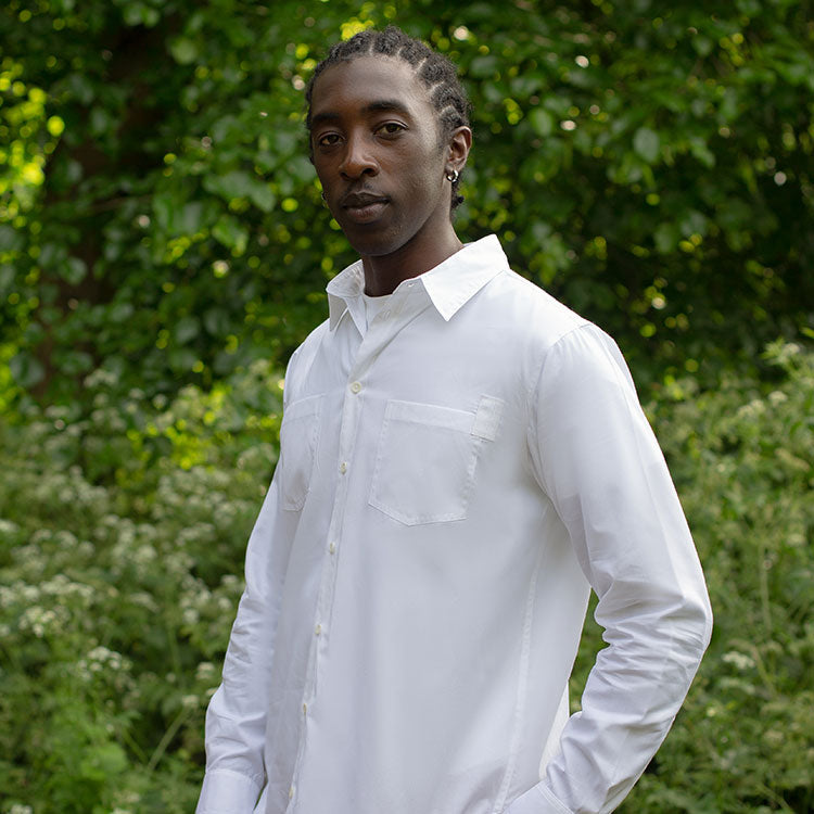 Male model stands with his hand in his pocket, wearing Saywood's Eddy Mens white cotton bamboo shirt, made in London, with patch pockets. He is surrounded by trees and greenery.
