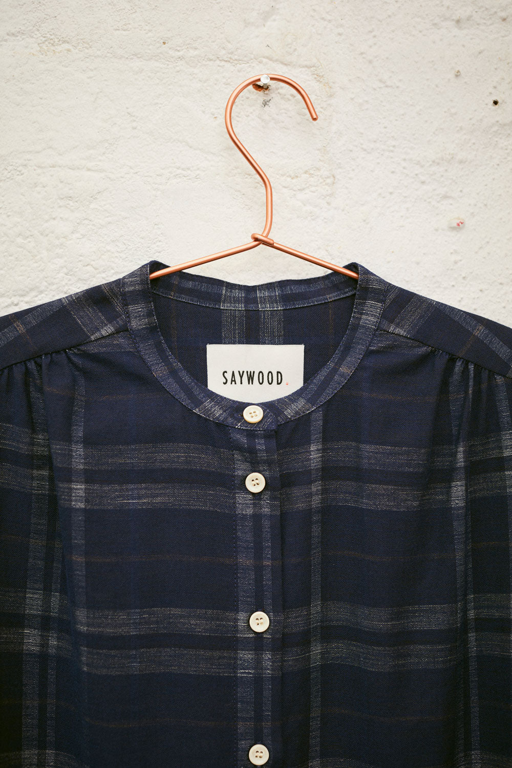 Close up of the Rosa Puff Sleeve Shirtdress in navy check, hanging on a hanger on a white wall. Only the neckline and shoulder gathers can be seen, with the Saywood label on the dress.