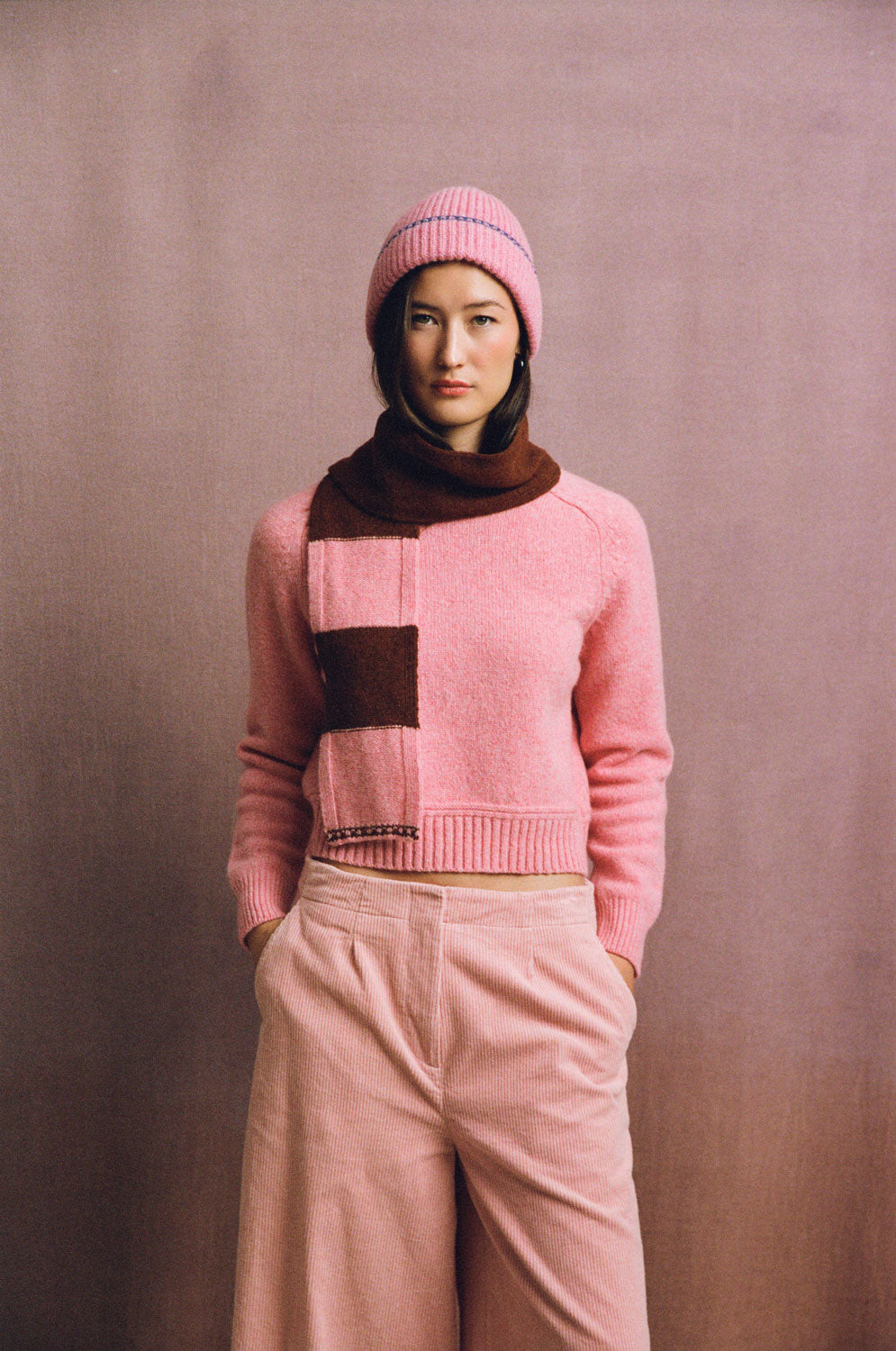 Front view of a model wearing a pink ribbed beanie with blue trim, a pink sweater and a pink and brown scarf.