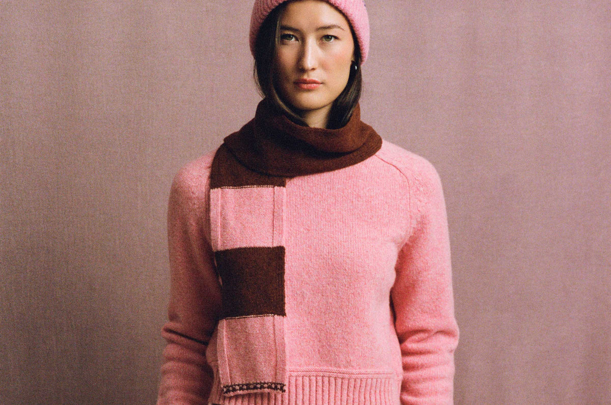 Pink Rosalind sweater with a ribbed hem, worn by a model with a pink knitted hat and rust-and-pink striped scarf against a muted pink background.