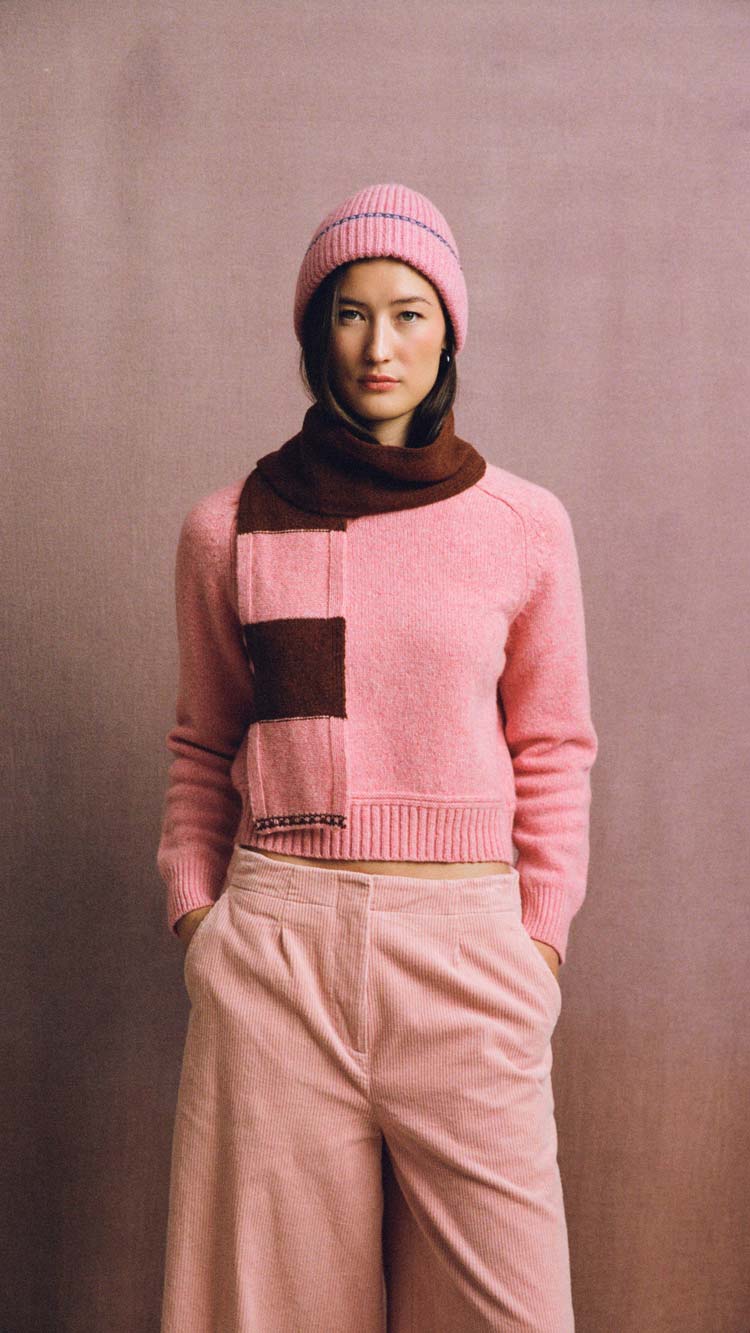 Pink Rosalind sweater with a ribbed hem, worn by a model with a pink knitted hat and rust-and-pink striped scarf against a muted pink background.