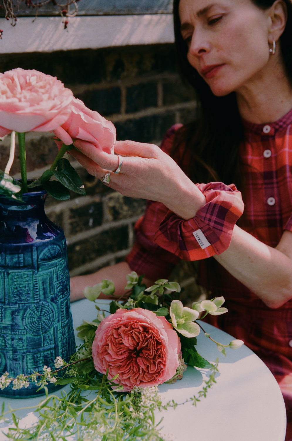 A woman sits at a blue garden table preparing pink flowers in a blue vintage vase. She is wearing Saywood's Rosa Puff Sleeve Shirtdress in coral red check with a matching scrunchie on her wrist.