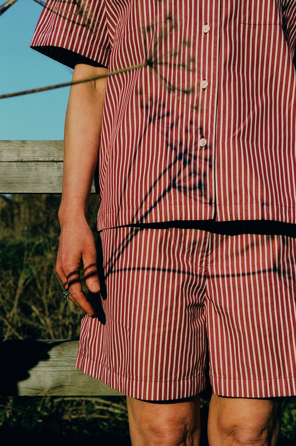 Close up of the upper body of a woman from her knees up. She has her hands by her side and is wearing Saywood's Frances Unisex Cuban Shirt and Bessie A-line Shorts in berry red stripe as a matching set. A stemmed flower is in front of her with shadows of the flowers playing on the shirt. Behind can be seen blue sky, grass and a wooden planked fence.