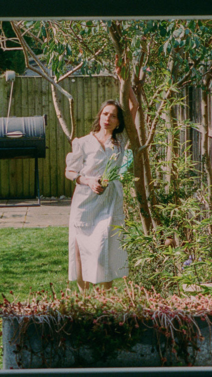 A woman is leaning, with one arm up, on a eucalyptus tree, holding a bunch of daffodils, in a garden. She is wearing Saywood's white pinstripe cotton Rosa Puff Sleeve Shirtdress, with ruffle trims on the bottom of the pocket edge. The image is shot through a window, with a window box on the window seal.