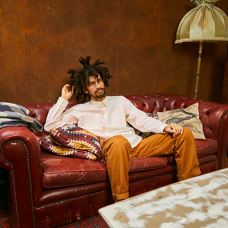 A man sits on a red leather sofa with cushions and a vintage lamp in the background. He wears Saywood's Eddy Men's Colourblock Shirt in Orange and Lemon pastel tones, made in London. Worn with tobacco trousers.