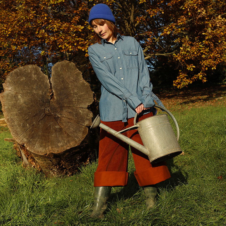 A woman stands in a grassy field, with a fallen tree in the background and Autumnal trees behind her. She is holding a watering can and wearing Saywood's Zadie Boyfriend Shirt in Japanese Denim with the Amelia Wide Leg Full Length Trousers in organic cotton caramel corduroy, styled rolled up, with green wellington boots and a blue beanie hat.