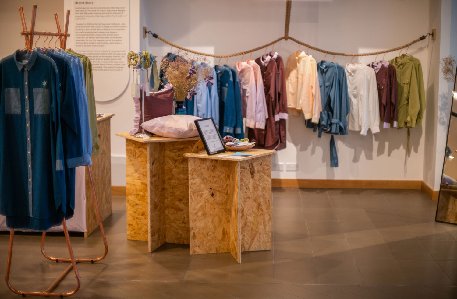 Saywood sustainable shirts and shirtdresses hanging on a rope rail in the background, the Japanese denim Etta Shirtdress in the foreground hanging on a copper rail, on Regent Street, London