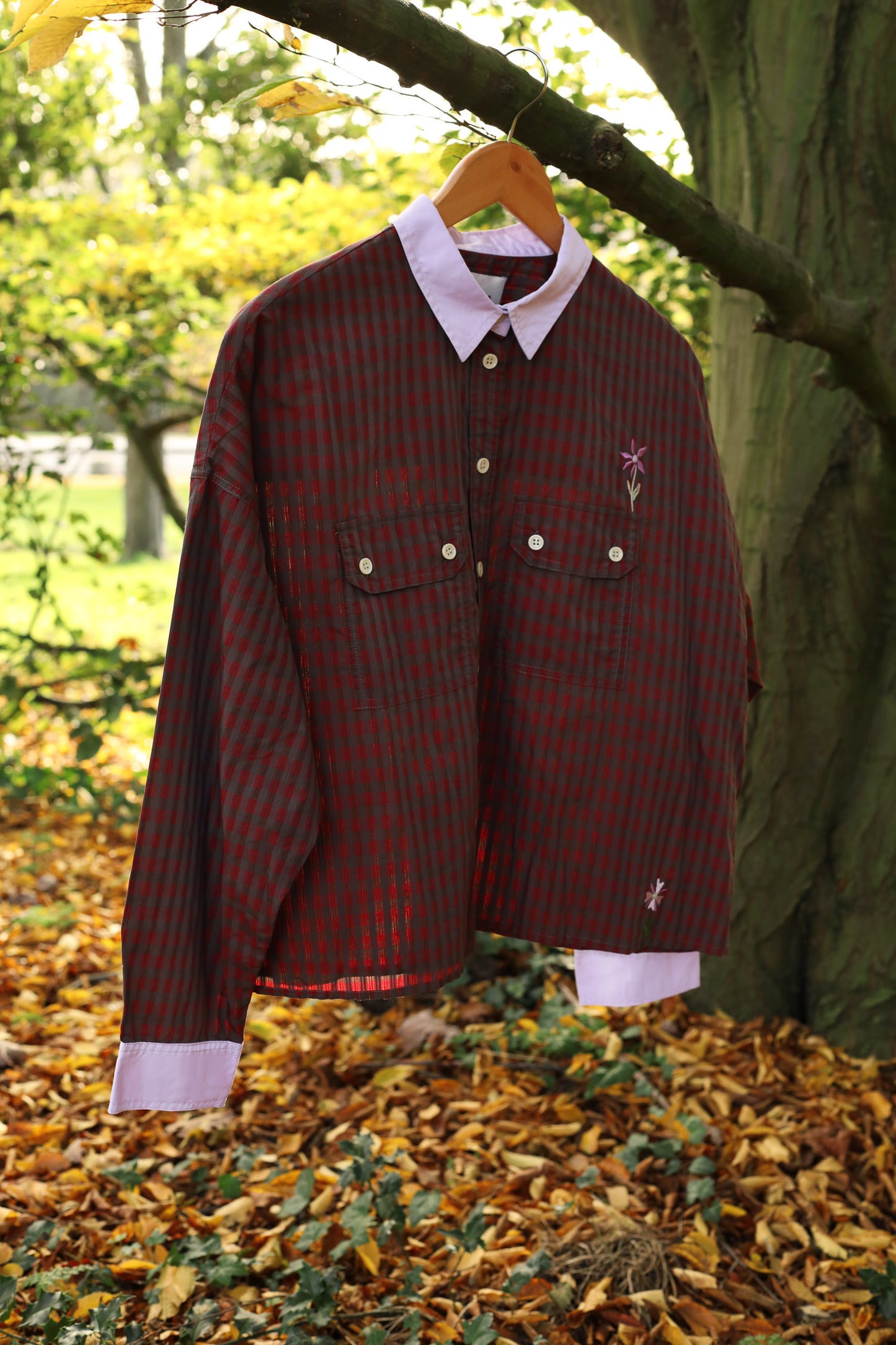 Women's red check shirt with lilac collar and cuffs hangs on a tree branch, with Autumnal leaves on the ground. The shirt has utility pockets and two embroidered flowers on. Made in Britain, by Saywood.