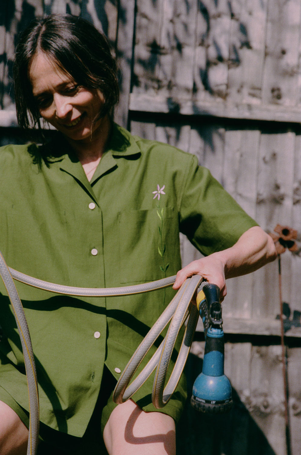 A woman is perched on a garden pot, holding a garden hose, with a wooden fence behind her. She is wearing the Frances Unisex Cuban Shirt in olive green, with short sleeves and the signature embroidered flower in lilac on the check pocket. Worn with the Bessie A-line Shorts in olive green as a matching twin set.