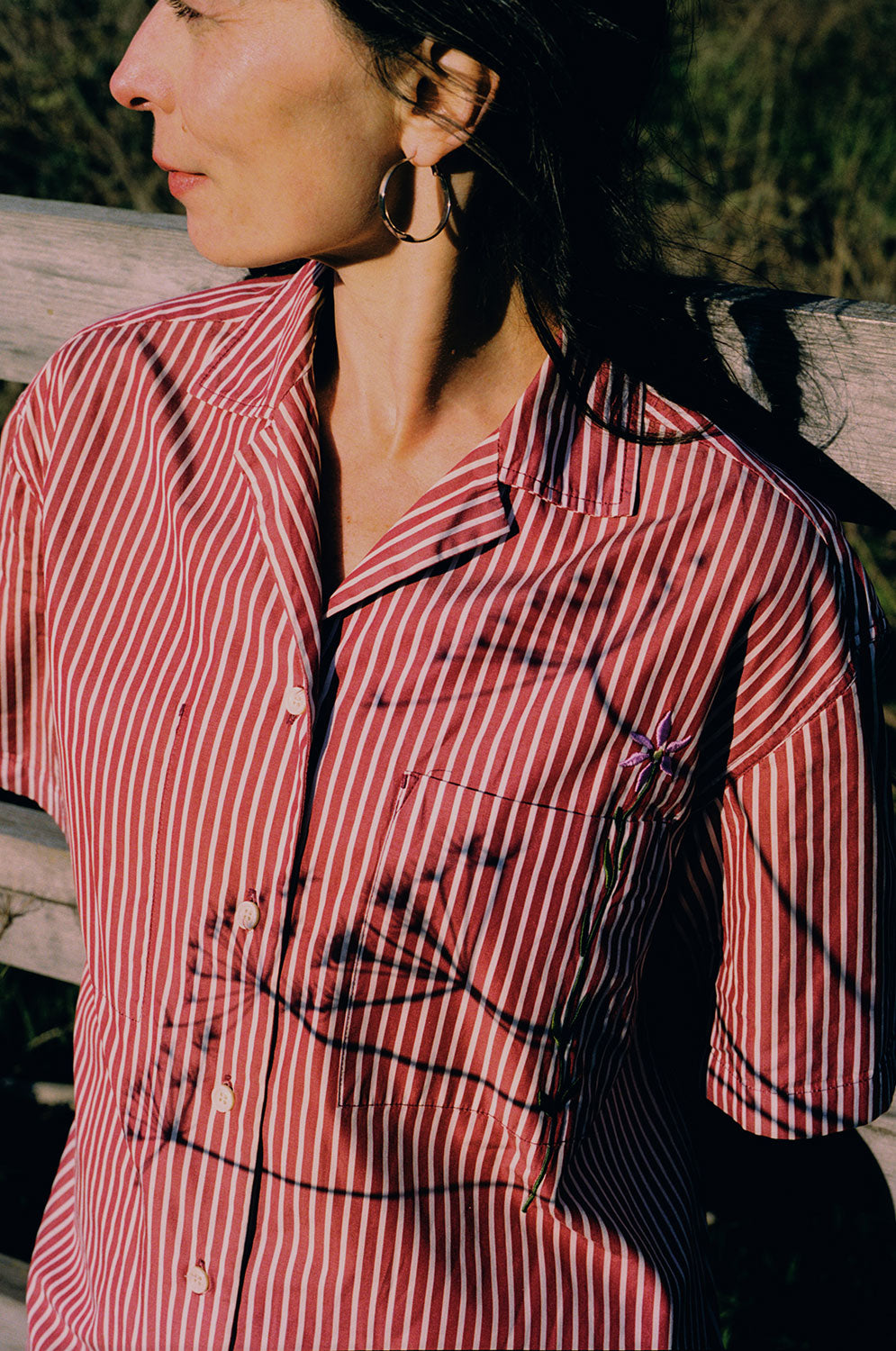 Close up of the Frances Unisex Cuban Shirt in red and white stripe. A woman is wearing the short sleeve shirt and the crop of the image shows the Saywood signature embroidered flower on the right chest pocket, and revere collar. Grass and a wooden fence can be seen behind her. She has her arms behind her back and is looking off to the side.