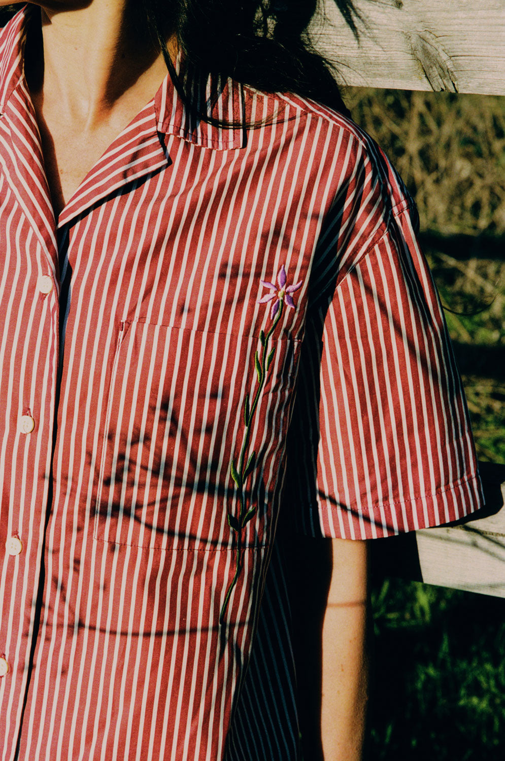 Close up of the Frances Unisex Cuban Shirt in red and white stripe. A woman is wearing the short sleeve shirt and the crop of the image shows the Saywood signature embroidered flower on the right chest pocket. Grass and a wooden fence can be seen behind her.