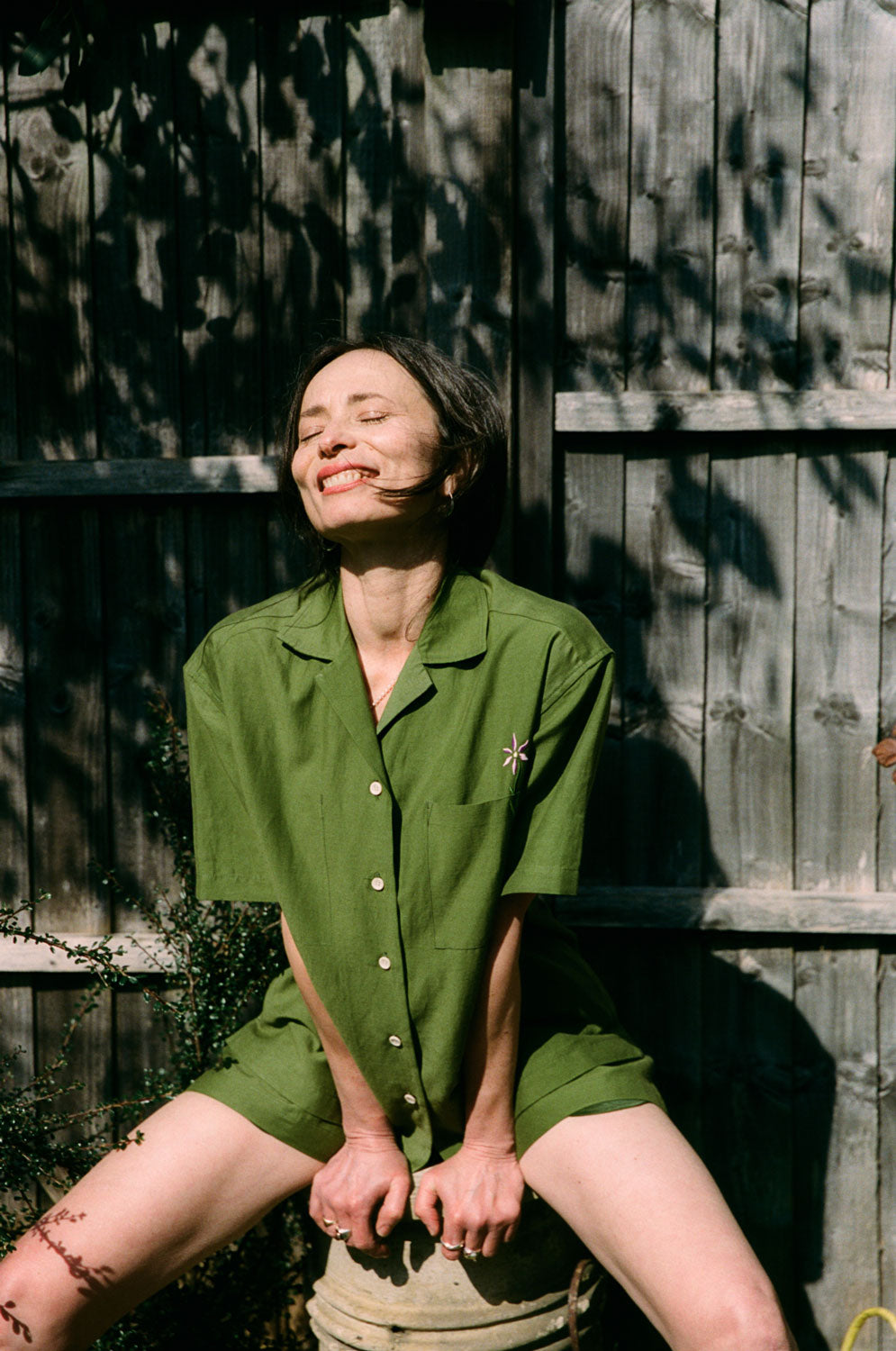 A woman is perched on a garden chimney pot, smiling with her eyes closed, basking in the sunlight. She is wearing the Frances Unisex Cuban Shirt in olive green, with short sleeves and the signature embroidered flower in lilac on the check pocket. Worn with the Bessie A-line Shorts in olive green as a matching twin set. She is leaning slightly forwards with her hands on the pot in front of her. A wooden fence is behind her with leafy shadows.
