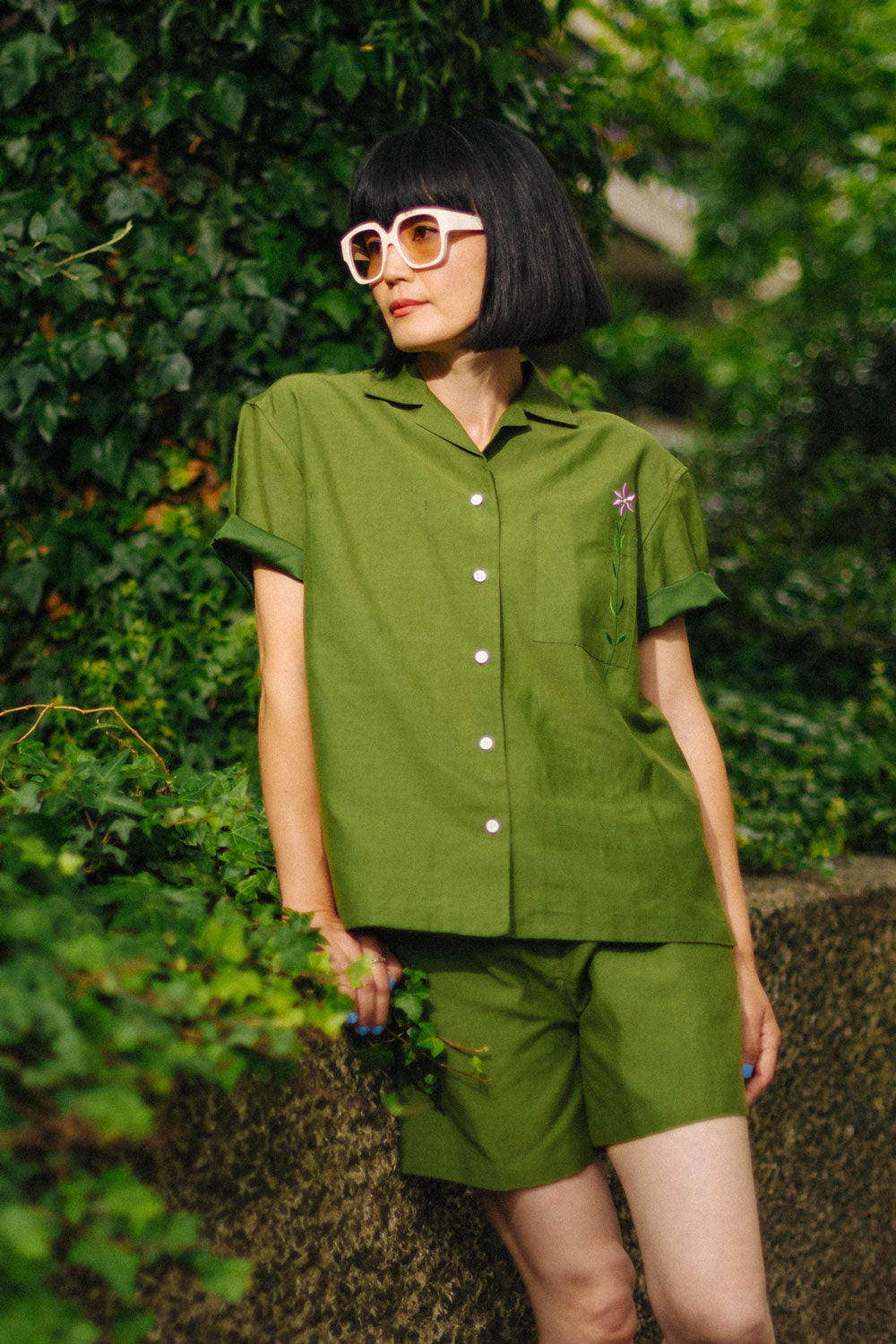 Woman wears the Saywood Frances Unisex Cuban Shirt, in olive green, with matching Bessie Aline Shorts. Made in London, the shirt is unisex, with short sleeve in a Cuban style, with an embroidered flower through the chest pocket. She wears the matching Bessie Shorts with pale pink sunglasses, and leans against a stone wall with green ivy behind her.