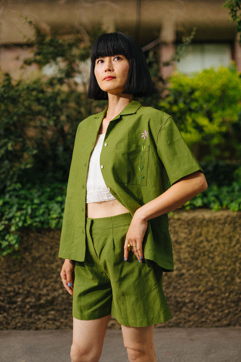 Woman wears the Saywood Frances Unisex Cuban Shirt, in olive green, with matching Bessie Aline Shorts. The shirt is worn open with a white crop top underneath. Made in London, the shirt is unisex, with short sleeve in a Cuban style, with an embroidered flower through the chest pocket. She wears the matching Bessie Shorts. She stands with one hand resting on the edge of the shorts pocket, in front of a stone wall with and greenery behind her.