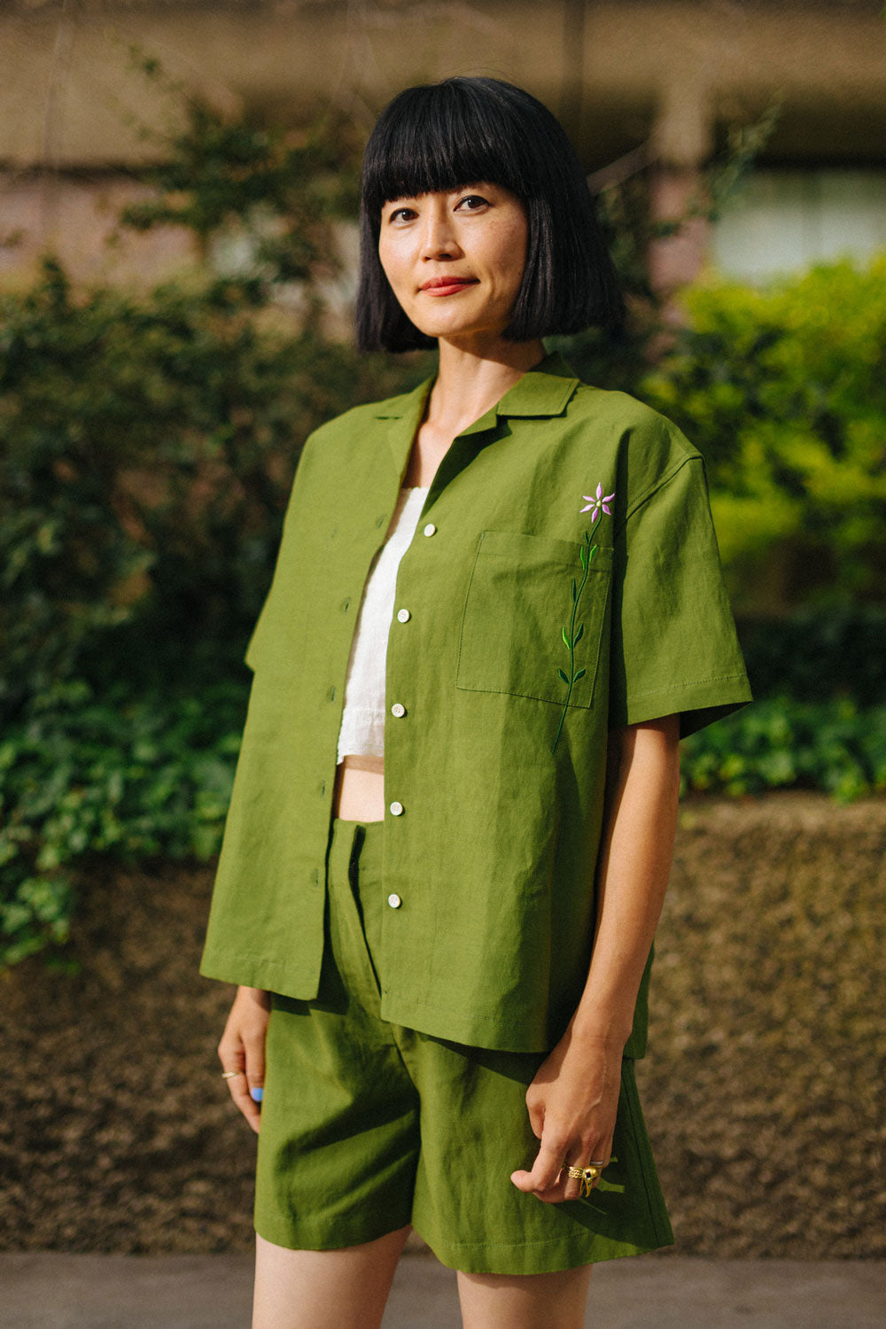 Woman wears the Saywood Frances Unisex Cuban Shirt, in olive green, with matching Bessie Aline Shorts. The shirt is worn open with a white crop top underneath. Made in London, the shirt is unisex, with short sleeve in a Cuban style, with an embroidered flower through the chest pocket. She wears the matching Bessie Shorts. She stands in front of a stone wall with and greenery behind her.