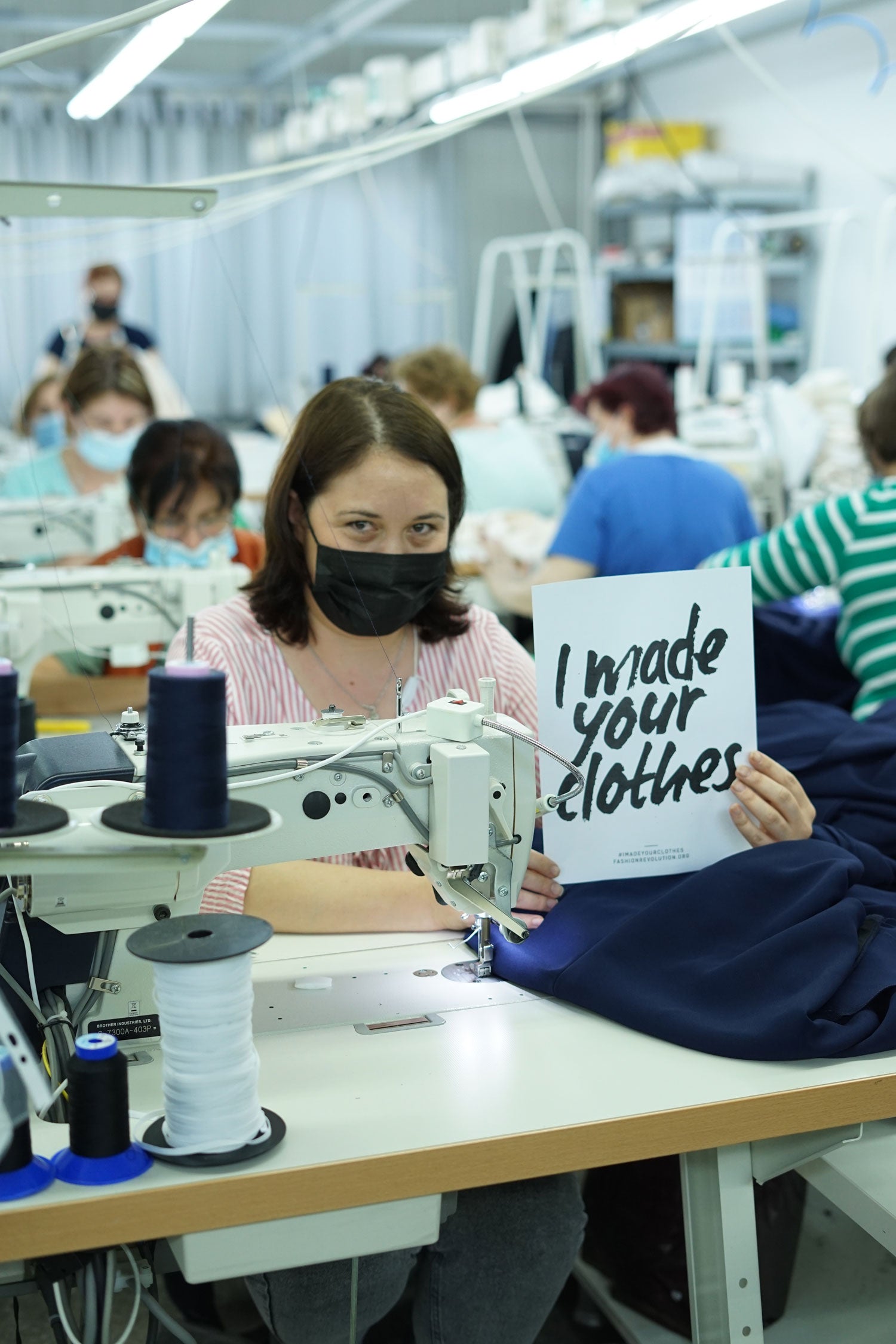 Machinist sits behind a sewing machine in a garment factory, wearing a mask, holding a sign that reads 'I made your clothes' Image is from our garment maker, Mantra, in Romania.
