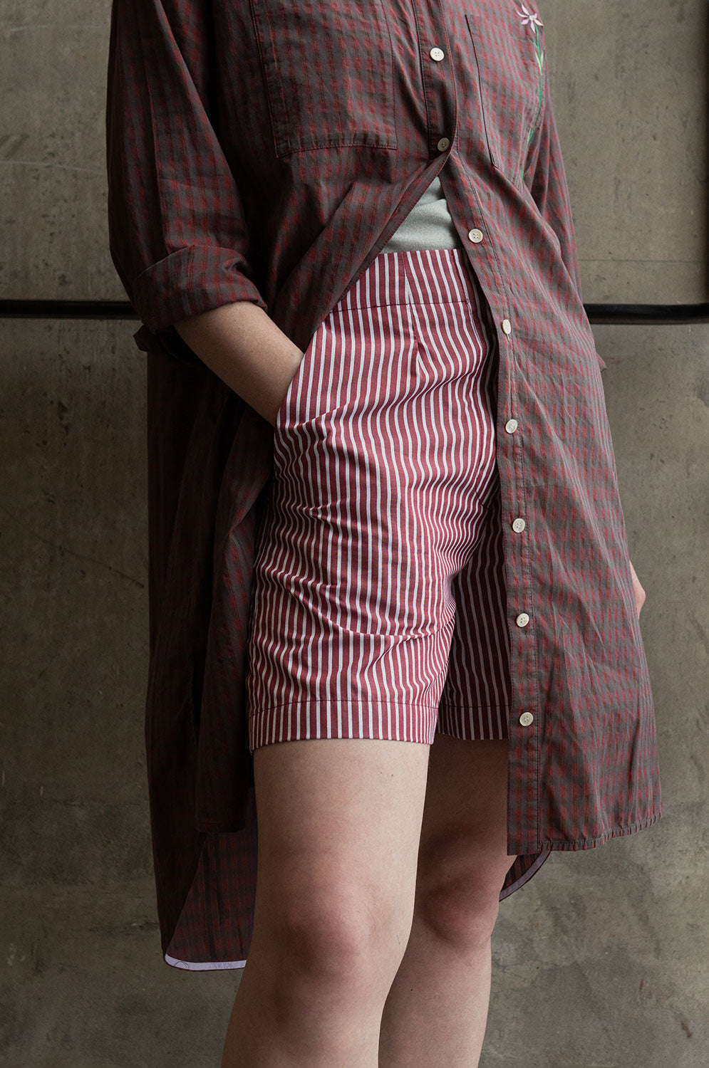 Close up of the lower body of a woman. She is wearing Saywood's Bessie A-line Shorts in berry red stripe, with the Etta Oversized Shirtdress in micro red check, half unbuttoned from the waist down. The woman has her hand in her shorts pocket.