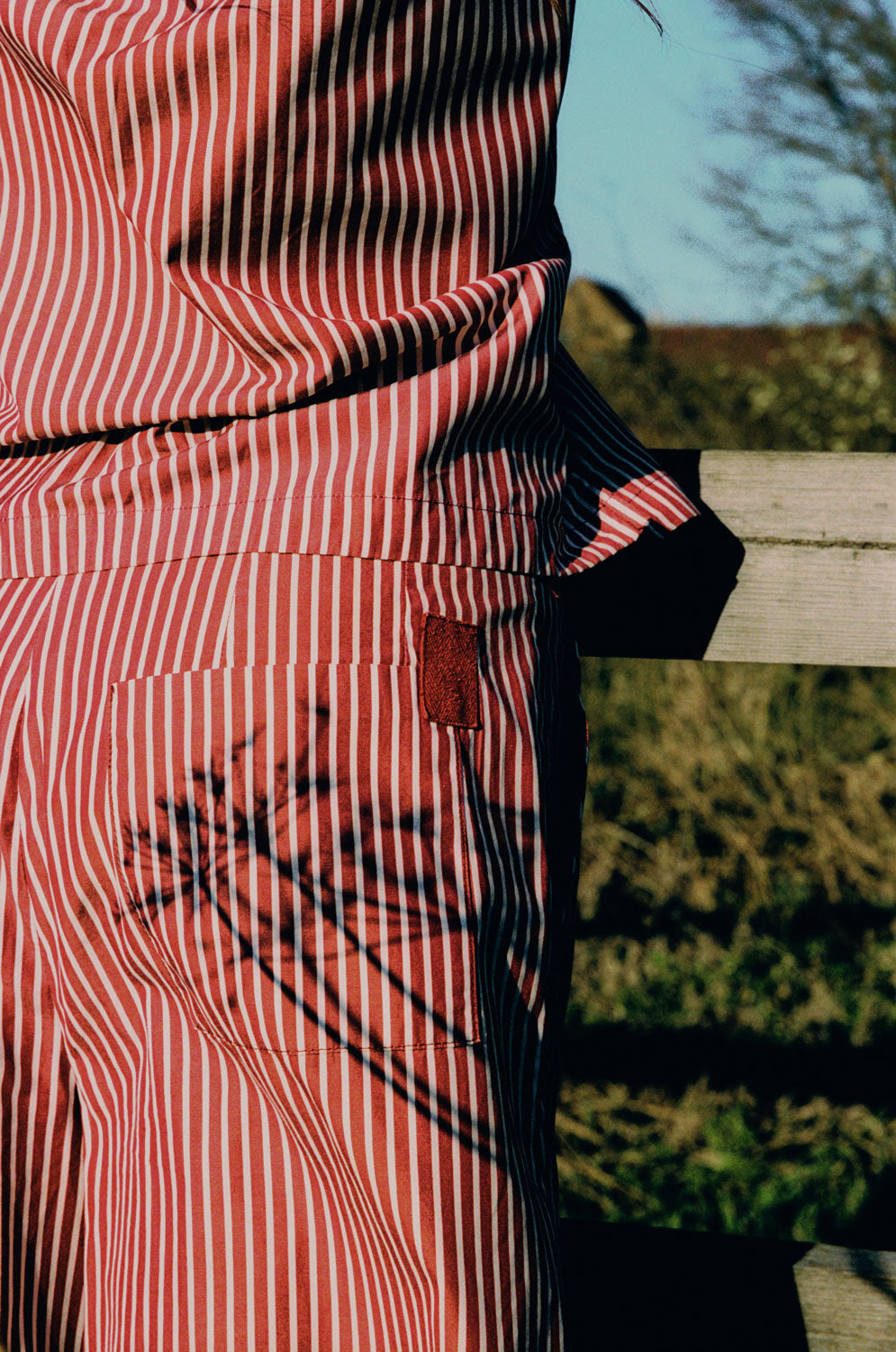 Close up detail shot of a woman wearing Saywood's Bessie A-line Shirts in berry red stripe. Shot from the back, the berry red cotton herringbone tape detail can be seen on the corner of the back patch pocket. The woman wears a matching shirt and is standing in front of a wooden fence with grass seen behind it.