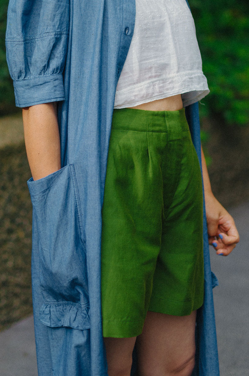 Close up of woman wearing Saywood's Bessie A-line Shorts in rich olive green. Worn with a white crop top, and Saywood's long line Rosa Dress in Japanese denim, worn open. Model has her hand in the dress pocket.