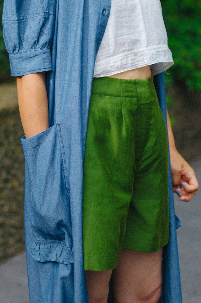 Close up of woman wearing Saywood's Bessie A-line Shorts in rich olive green. Worn with a white crop top, and Saywood's long line Rosa Dress in japanese denim, worn open. Model has her hand in the dress pocket.