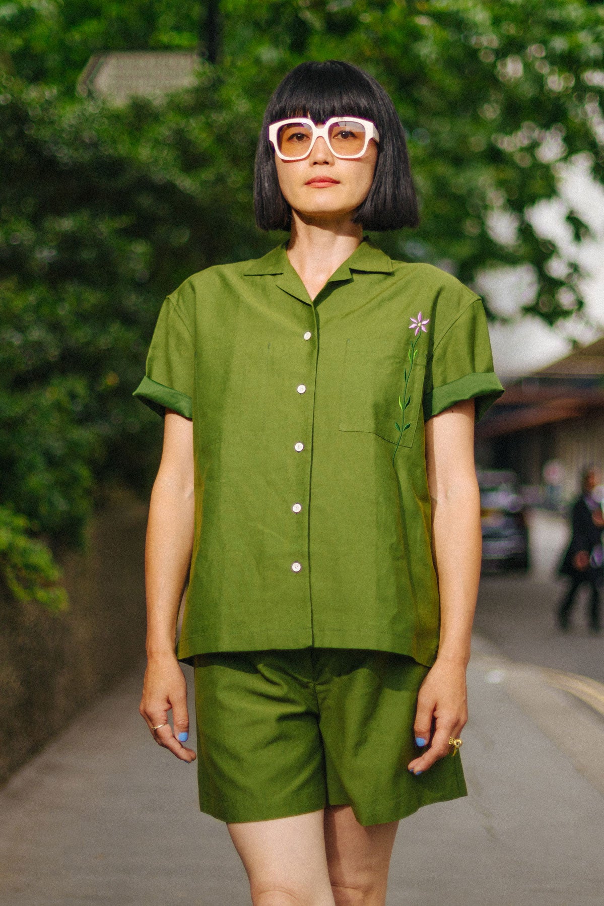 Woman stands straight on to the camera with one leg crossed in front of the other. She wears Saywood's Frances Unisex Cuban Shirt in olive green, with embroidered flower on the chest pocket, worn with the matching Bessie shorts in the olive green, and pale pink sunglasses. She stands on a street with greenery and concrete in the background.