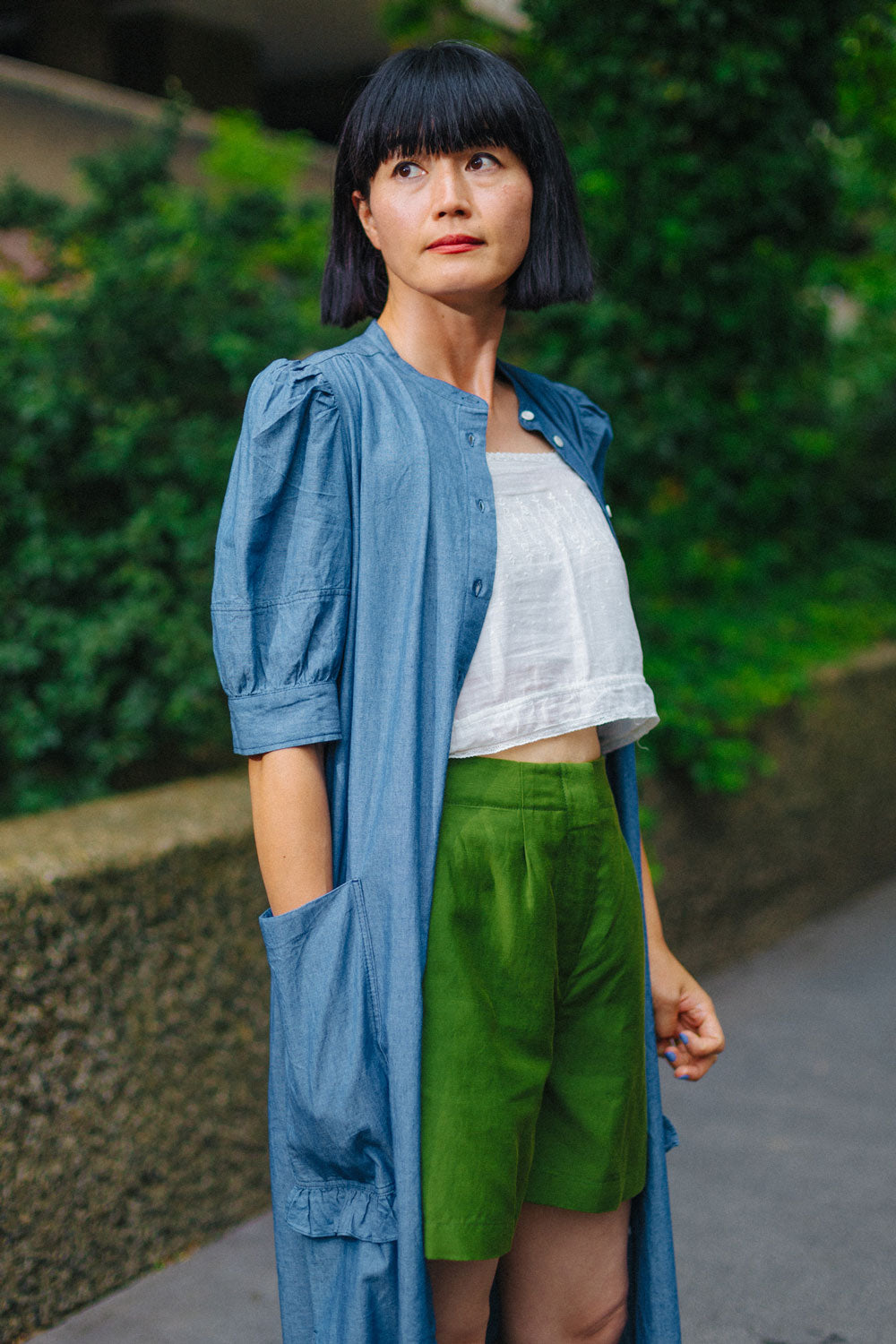 Woman stands side on to the camera, wearing Saywood's Bessie A-line Shorts in rich olive green. Worn with a white crop top, and Saywood's long line Rosa Dress in japanese denim, worn open. Model has her hand in the dress pocket, and greenery and a stone wall can be seen in the background..