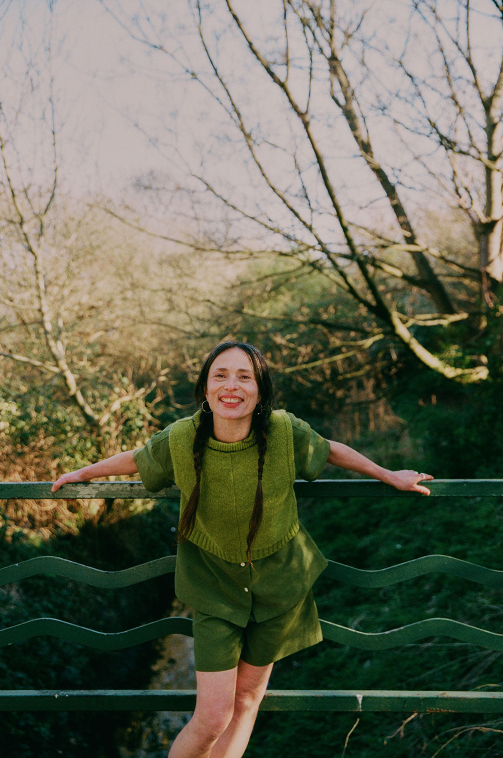 A woman is perched on a garden pot, holding a garden hose, with a wooden fence behind her, with leafy shadows on the fence. She is wearing the Frances Unisex Cuban Shirt in olive green, with short sleeves and the signature embroidered flower in lilac on the check pocket. Worn with the Bessie A-line Shorts in olive green as a matching twin set. She is holding a curled up garden hose, with one leg up on the garden chimney pot.