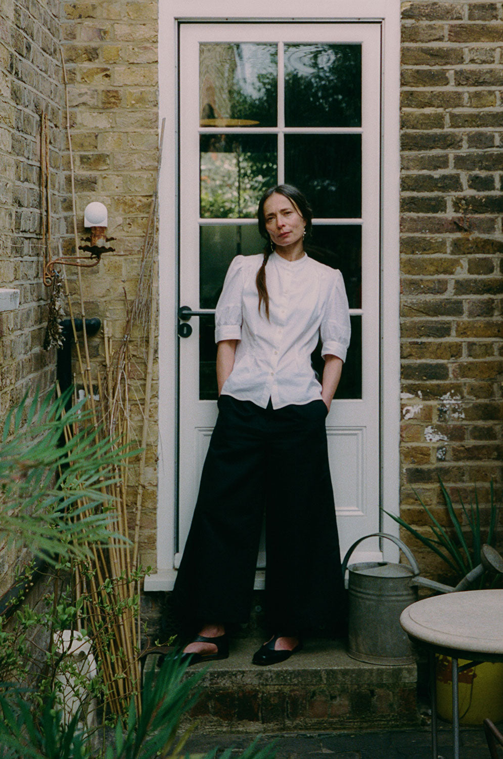 A woman stands in front of a back door in white with glass window panes. She is standing in a garden side alley, with plants in front of her and a small table. Her hands are in her trouser pockets. She wears Saywood's Amelia Wide Leg Culotte Trousers in black organic cotton and linen, with the Joni Puff Sleeve Blouse in white Supima cotton and bamboo viscose, with black open shoes.