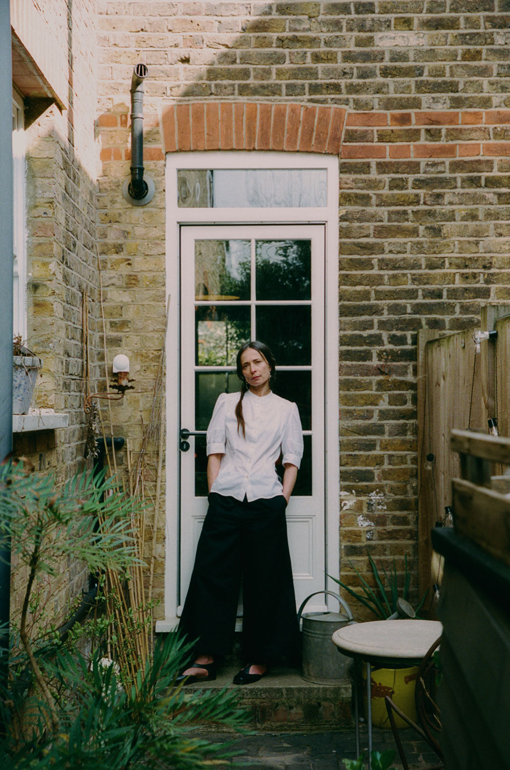 A woman stands in front of a back door in white with glass window panes. She is standing in a garden side alley, with plants in front of her and a small table. Her hands are in her trouser pockets. She wears Saywood's Amelia Wide Leg Culotte Trousers in black organic cotton and linen, with the Joni Puff Sleeve Blouse in white Supima cotton and bamboo viscose, with black open shoes.