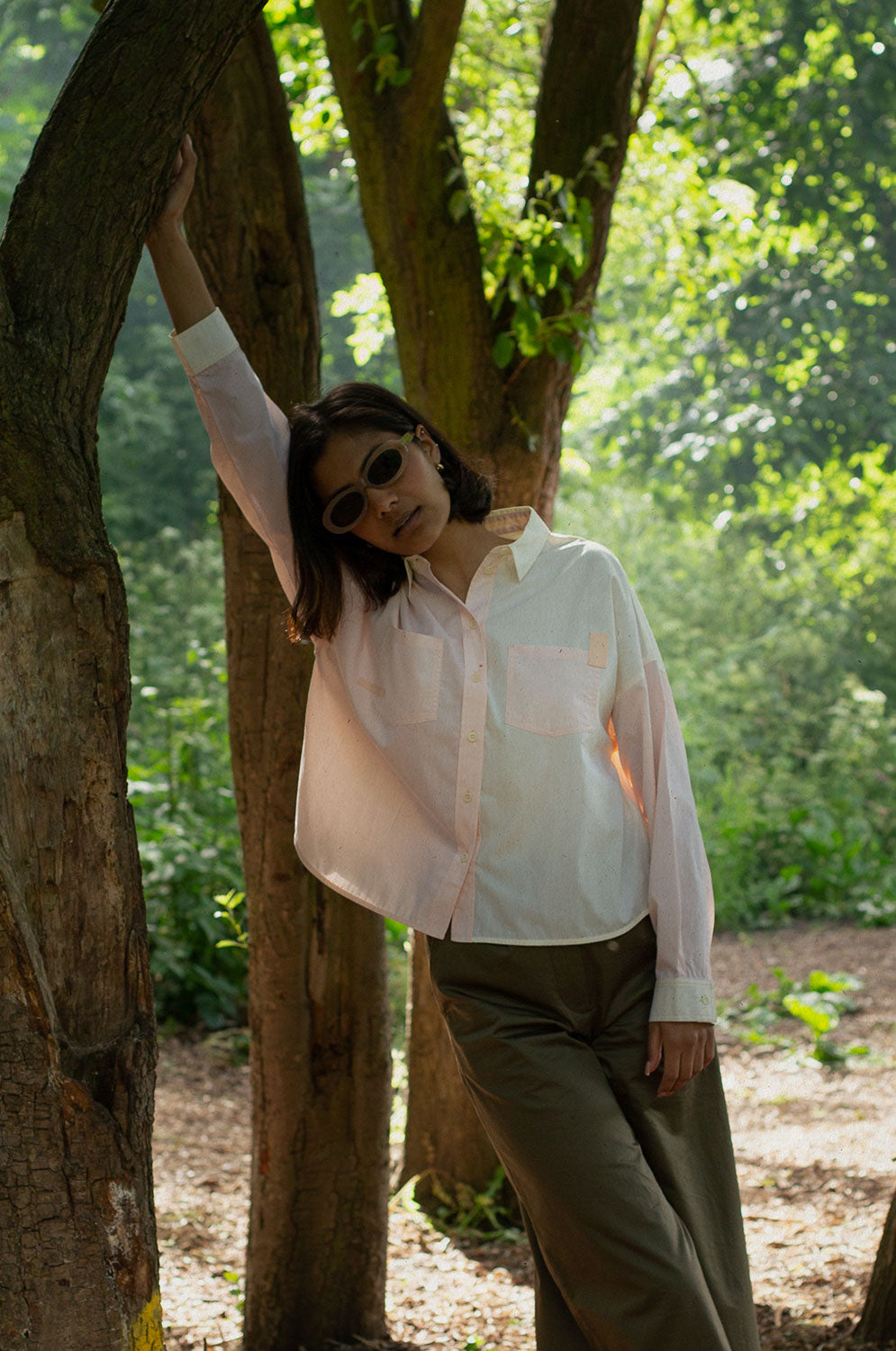 A woman stands, with one arm up leaning against the trunk of a tree. Trees and greenery can be seen behind her. She is wearing Saywood's Lela Colourblock Boxy shirt in lemon and orange vibrant pastel, with the Amelia Wide Leg Trousers in khaki. She has sunglasses on.