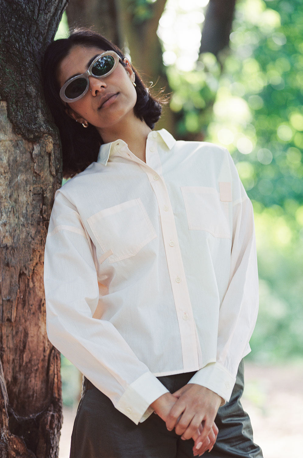 A woman stands, leaning on a tree. She is wearing Saywood's Lela Colourblock Boxy shirt in lemon and orange vibrant pastel. Worn with the Amelia Wide Leg Trousers in khaki cotton. She has sunglasses on.