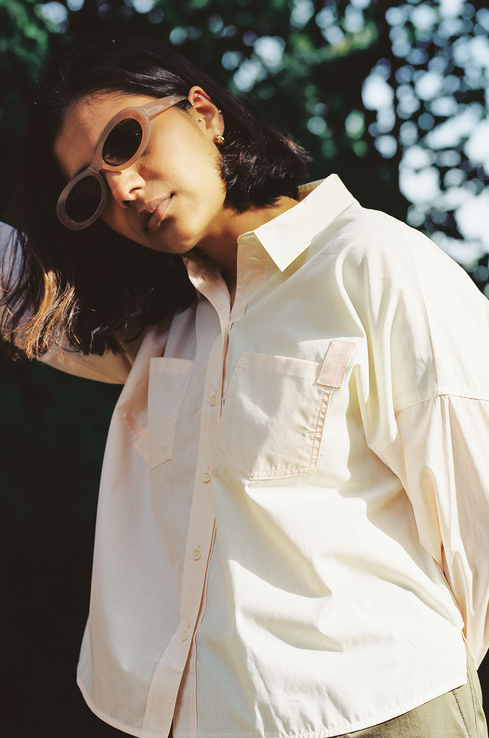 A woman stands, with one arm up leaning. Trees can be seen behind her. She is wearing Saywood's Lela Colourblock Boxy shirt in lemon and orange vibrant pastel. The patch pocket detail with cotton herringbone tape can be seen. She has sunglasses on.