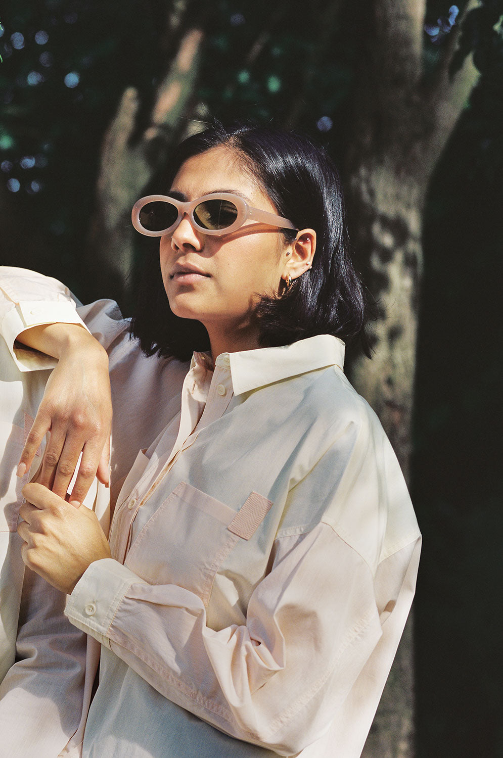 A woman stands, with one arm up leaning on someone's shoulder. Trees can be seen behind her. She is wearing Saywood's Lela Colourblock Boxy shirt in lemon and orange vibrant pastel. The patch pocket detail with cotton herringbone tape can be seen. She has sunglasses on.
